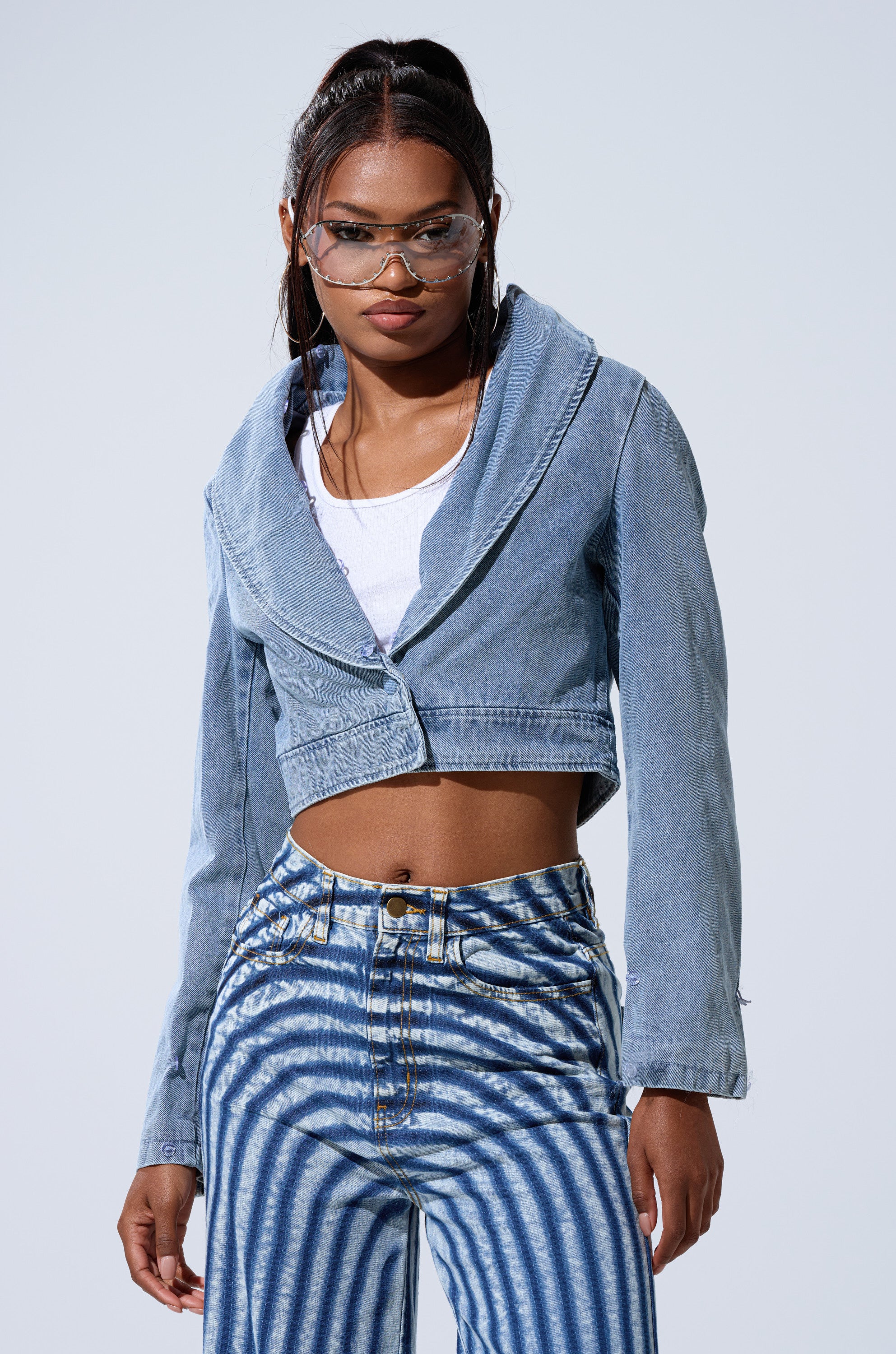 A woman wearing clear glasses, a white top, high-waisted blue and white striped pants, and the GISELE CROP DENIM BLAZER stands against a plain light background.