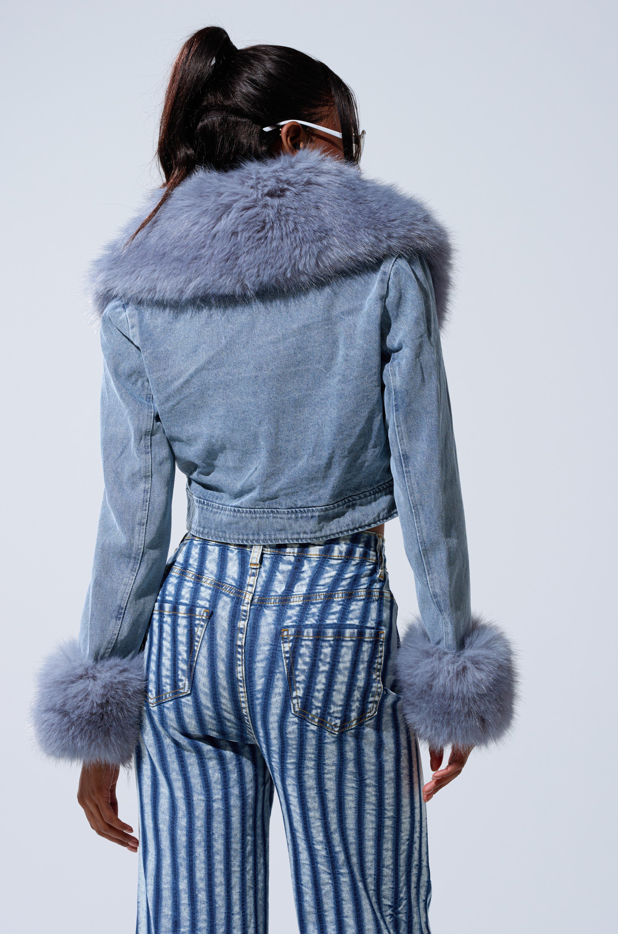 A person facing away wears the GISELE CROP DENIM BLAZER featuring a large gray faux fur collar and cuffs in Bratz Doll style, paired with high-waisted blue-and-white striped jeans. Hair is tied back in a ponytail.