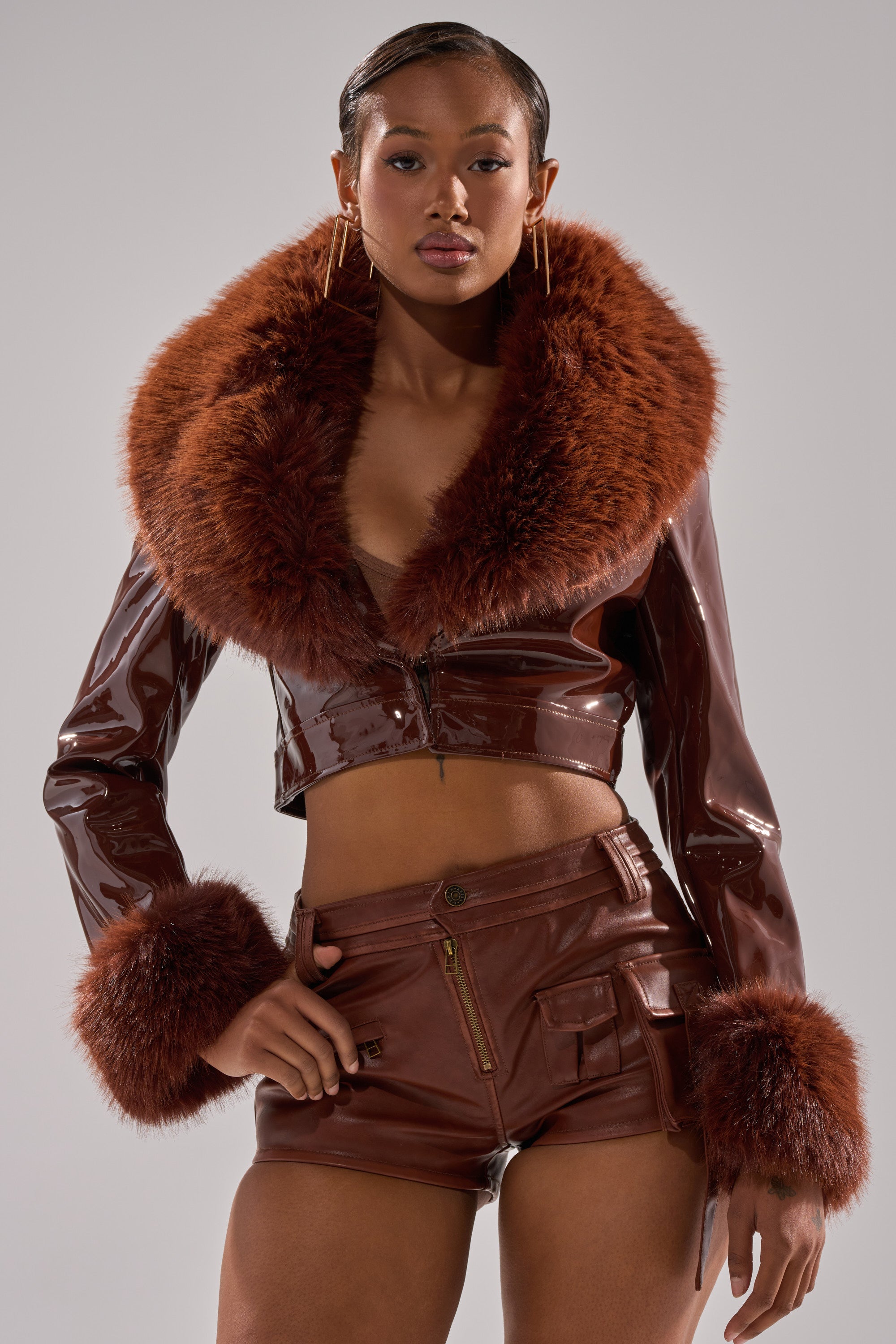 A woman wears the GISELE FUR TRIM MOTO IN PATENT—a shiny brown faux patent leather cropped jacket with bold faux fur collar and cuffs—paired with matching high-waisted shorts, standing confidently against a plain gray background.