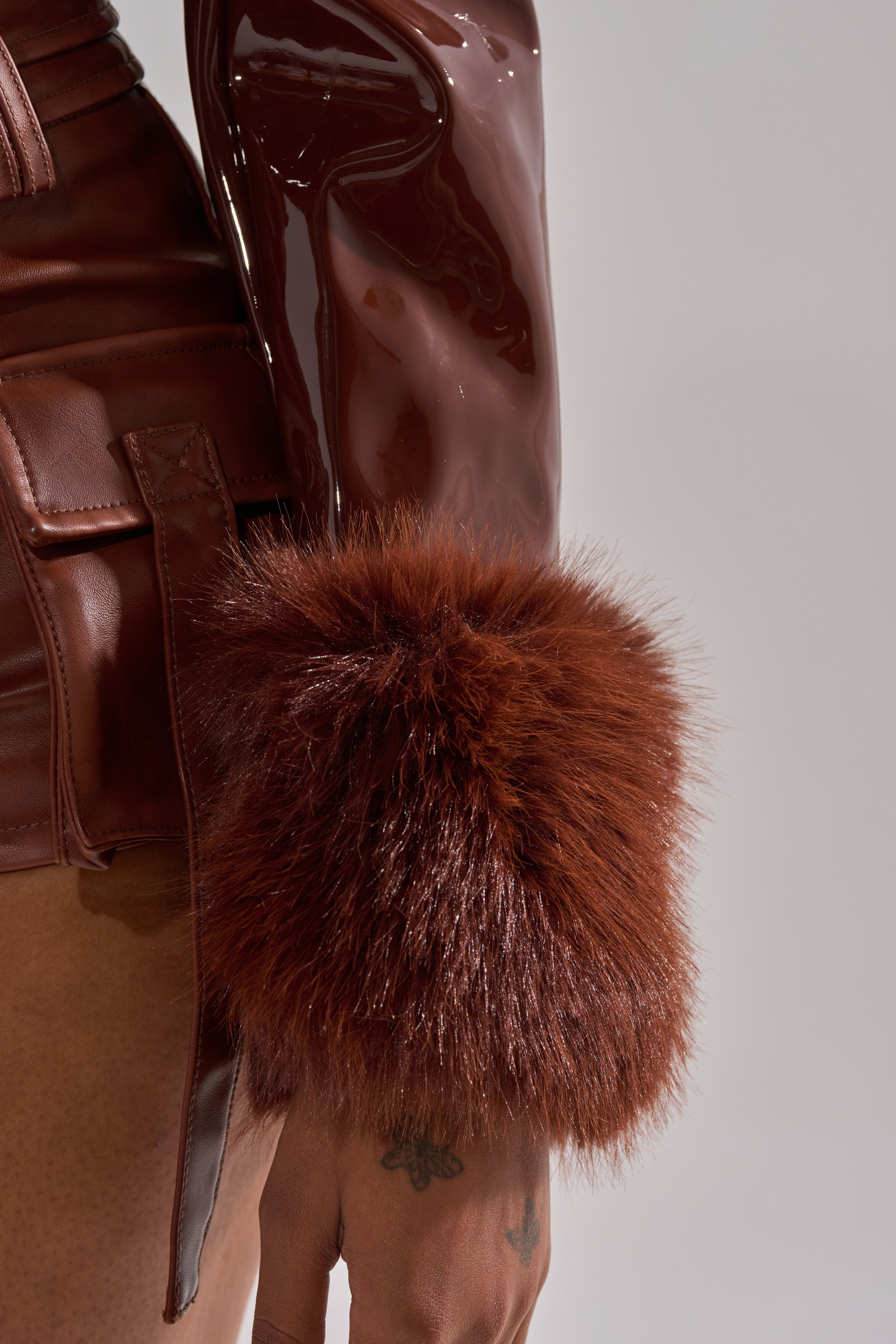 A close-up shows a person wearing the GISELE FUR TRIM MOTO IN PATENT, a glossy brown faux patent leather jacket with shiny sleeves and a large, fluffy fur cuff. A small tattoo is visible on the hand below the cuff.