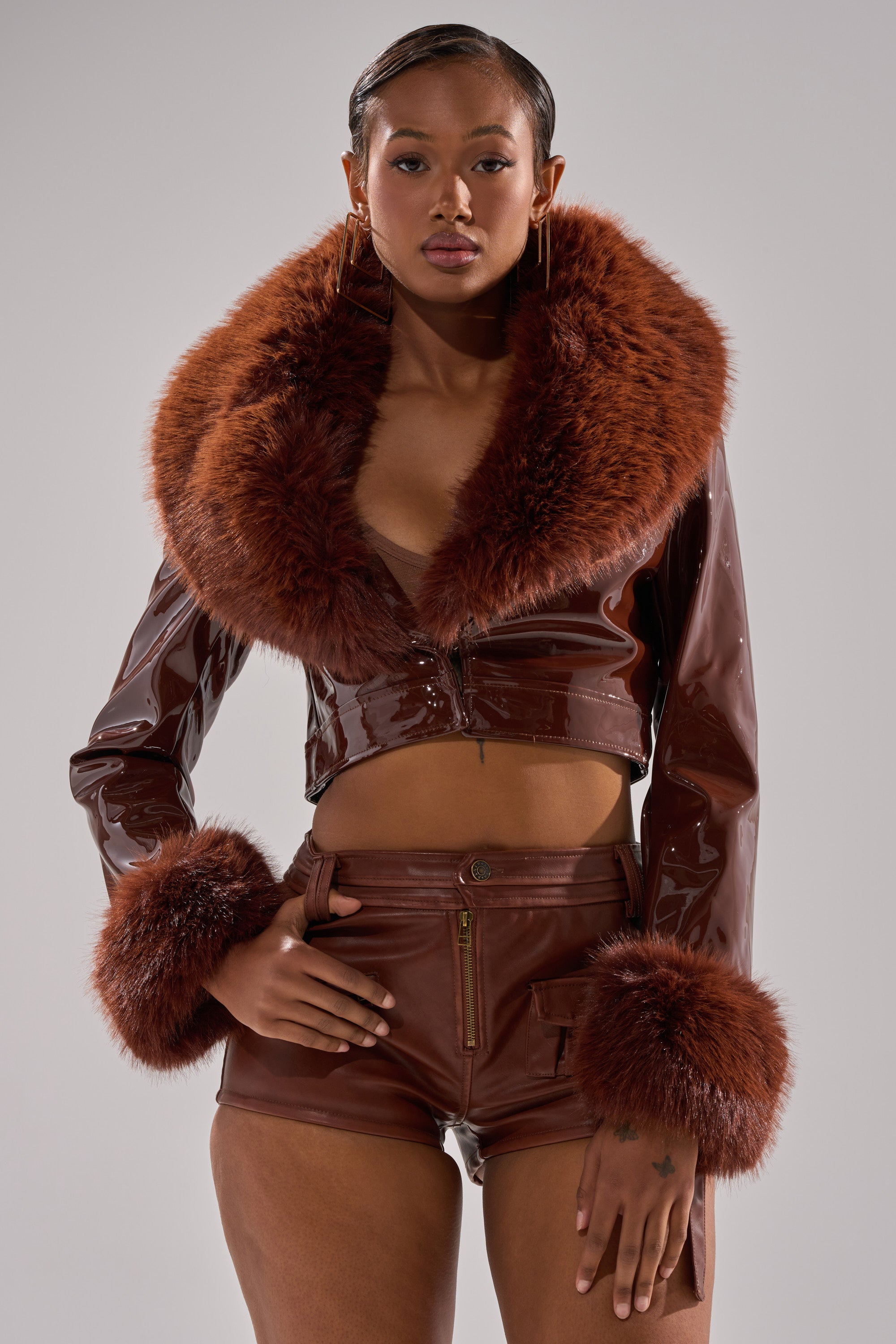 A woman poses confidently in the GISELE FUR TRIM MOTO IN PATENT, a glossy brown faux patent leather cropped jacket with large faux fur collar and cuffs, paired with high-waisted brown shorts against a plain light grey background.