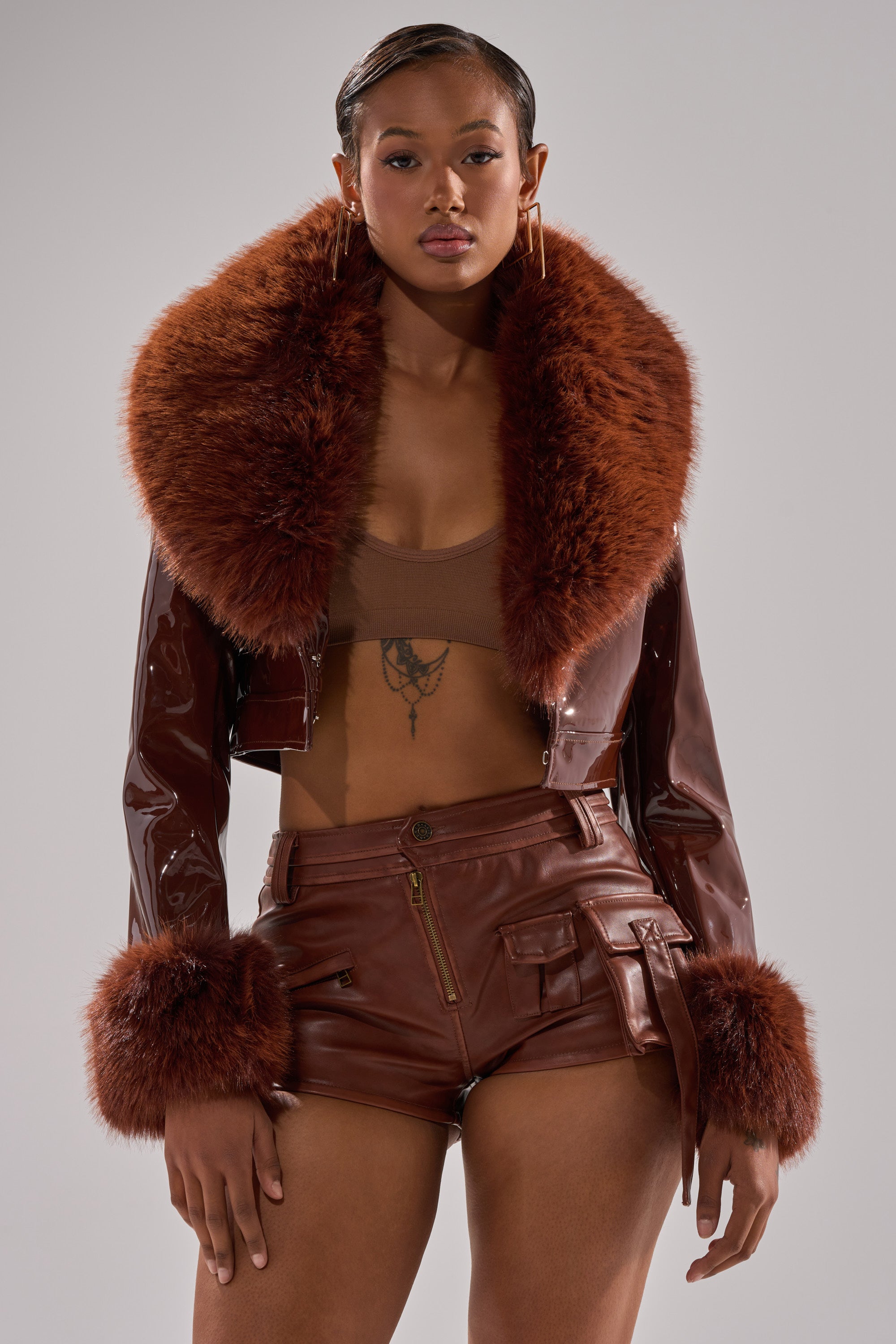 A woman faces forward wearing the GISELE FUR TRIM MOTO IN PATENT jacket, featuring a brown cropped design with a large, fluffy collar and cuffs, paired with matching high-waisted leather shorts and a brown crop top.