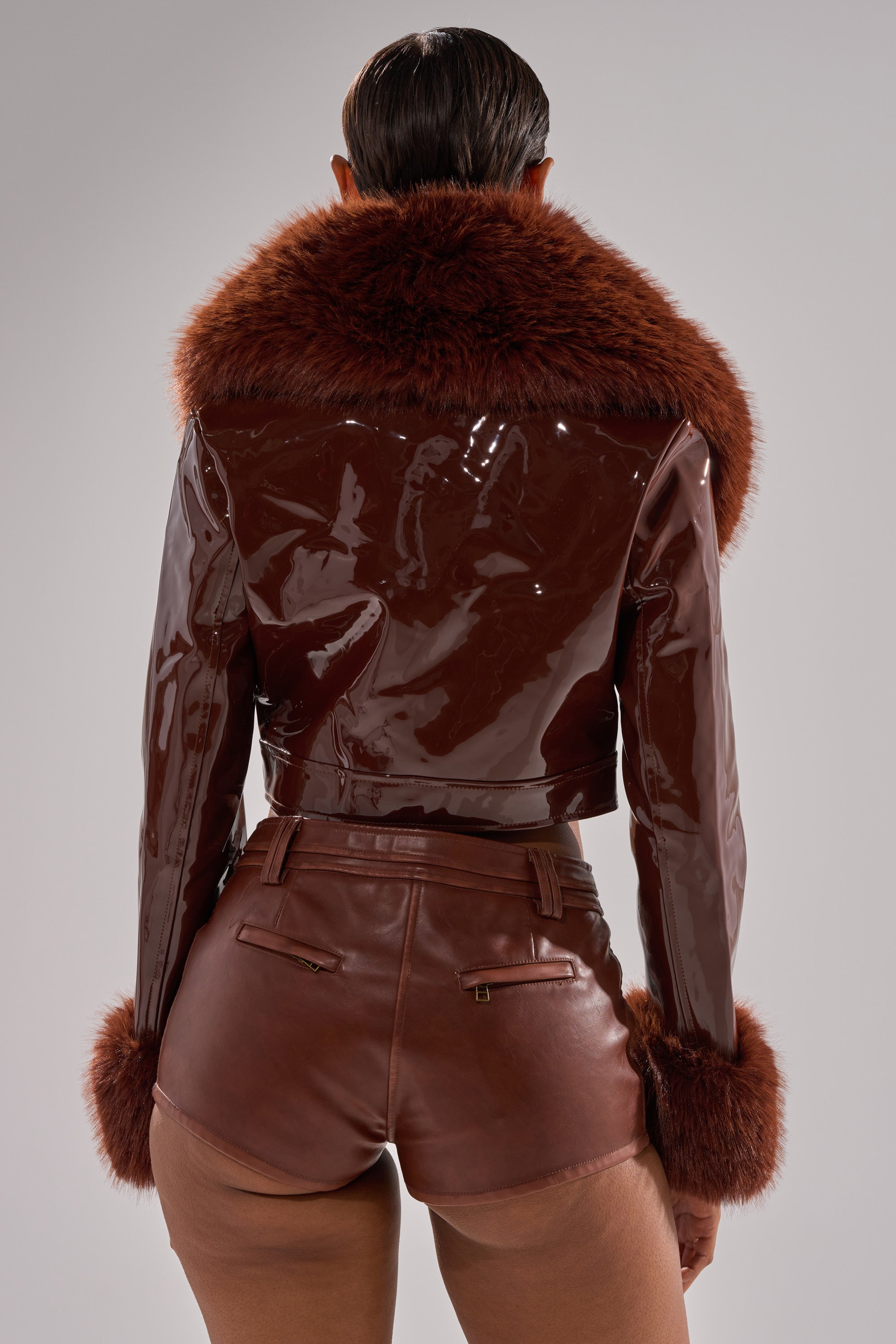 A person faces away, wearing the GISELE FUR TRIM MOTO IN PATENT—a glossy faux patent leather jacket with wide faux fur collar and cuffs—paired with high-waisted brown leather shorts against a plain, light background.