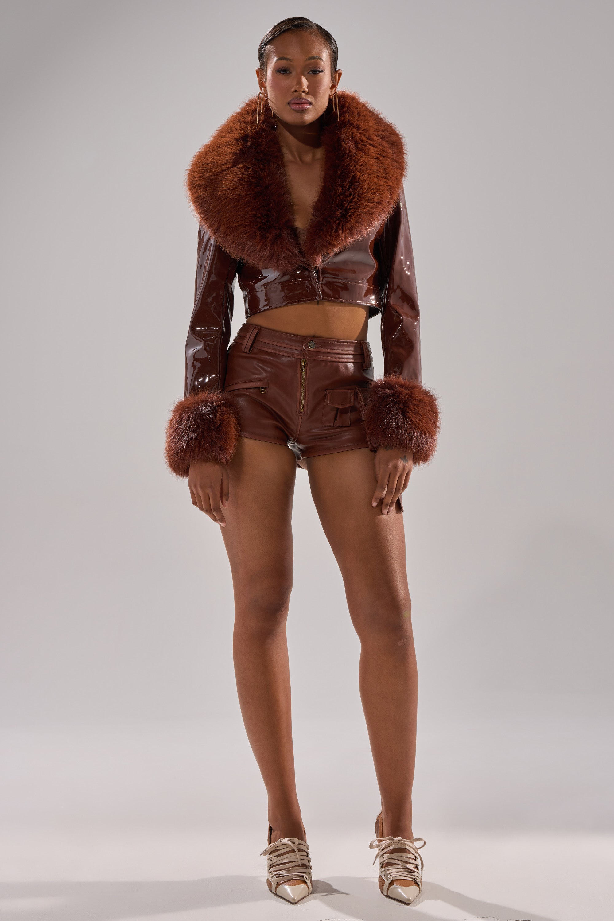 A woman poses confidently in a studio, wearing the GISELE FUR TRIM MOTO IN PATENT jacket with faux fur collar and cuffs, brown leather shorts, and gold heels against a plain gray background.