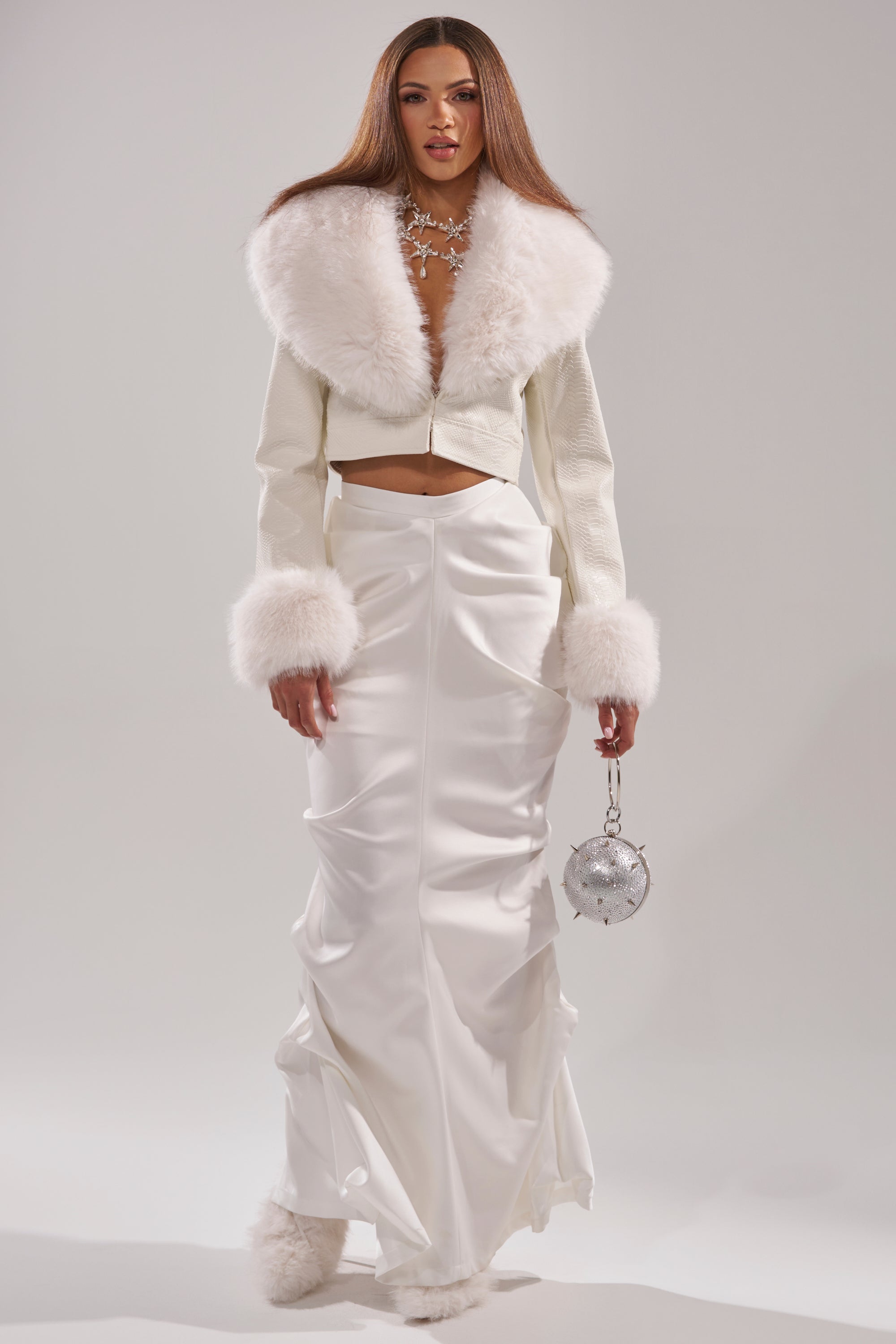 A woman with straight brown hair stands against a plain background wearing the GISELE FUR TRIM MOTO IN WHITE CROC jacket, a long white satin skirt, fluffy white croc shoes, and holding a small silver spherical bag.