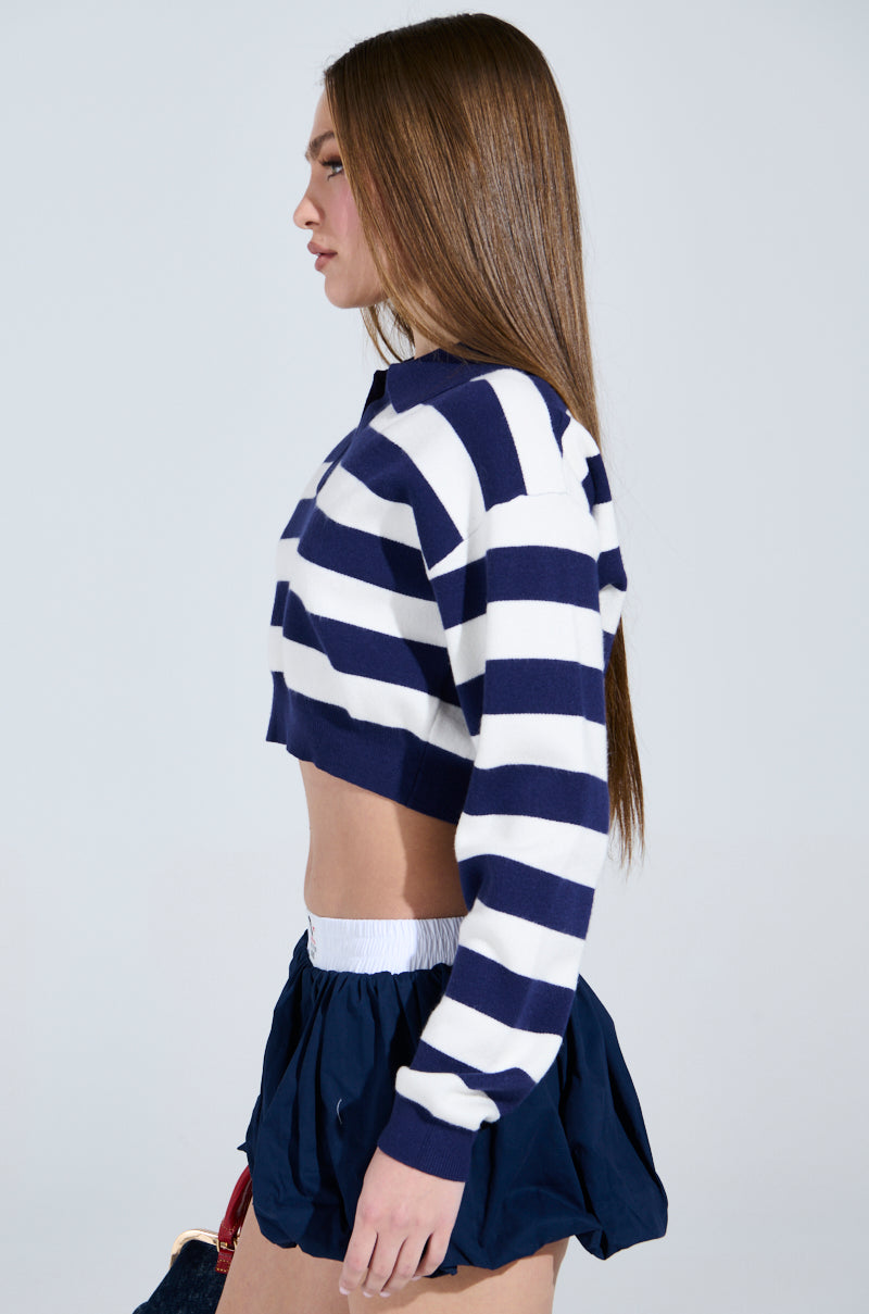 A person with long brown hair is wearing the CHIC ALWAYS SWEATER in navy and white rugby stripes, paired with a navy skirt with a white waistband, standing in profile against a plain background.