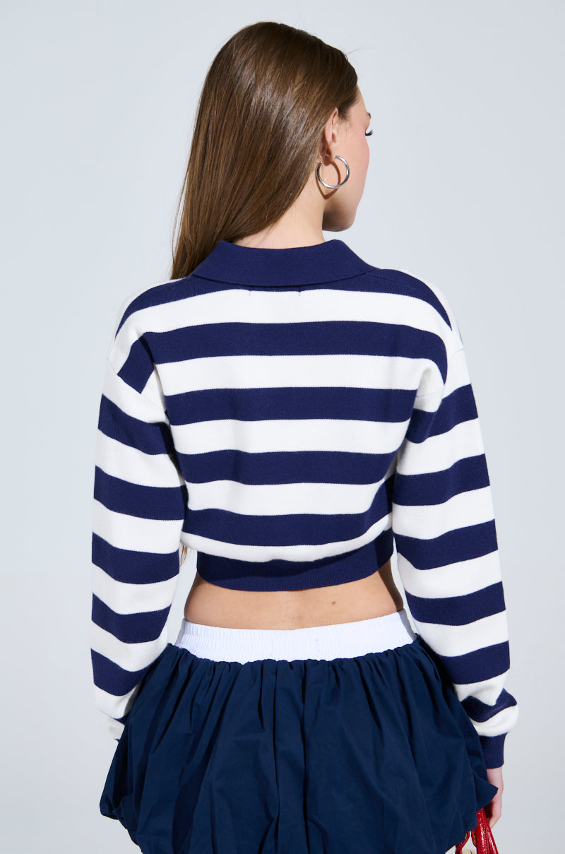 A woman with long brown hair and hoop earrings wears a CHIC ALWAYS SWEATER in rugby stripes. She pairs it with a navy skirt with a white waistband, standing against a plain backdrop, facing away from the camera.