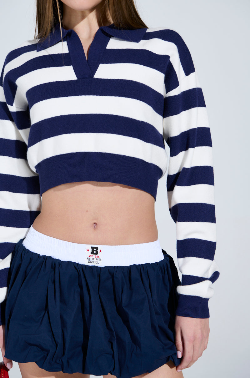 Someone wears the CHIC ALWAYS SWEATER in navy and white, featuring a chic rugby stripe pattern, paired with a dark blue skirt with an elastic waistband. Only the torso and part of the arms are visible against a plain background.