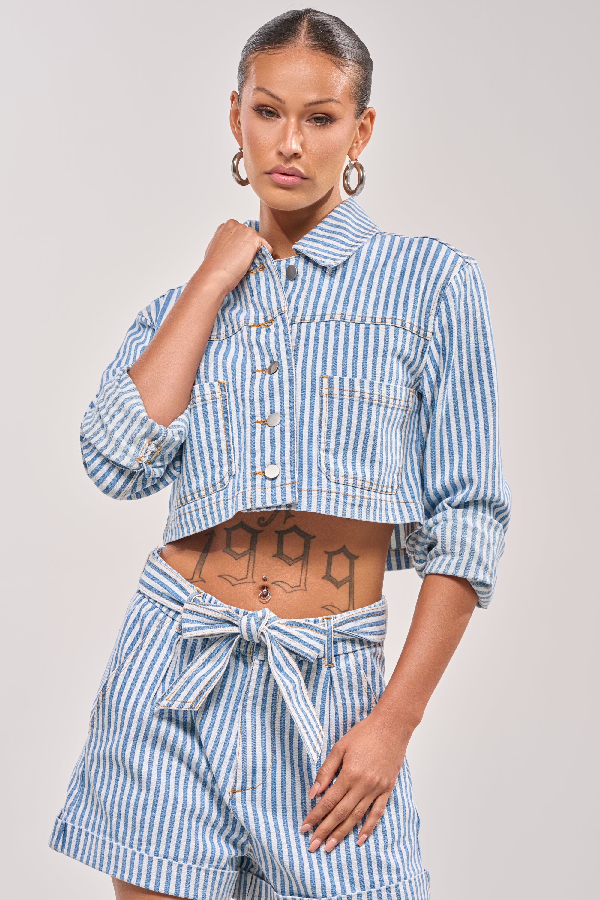 She wears the VACA VIBES BUTTON DOWN, a blue and white vertically striped, cropped boxy shirt with matching high-waisted shorts, hoop earrings, and pulled-back hair, showing off her "1999" tattoo. 0
