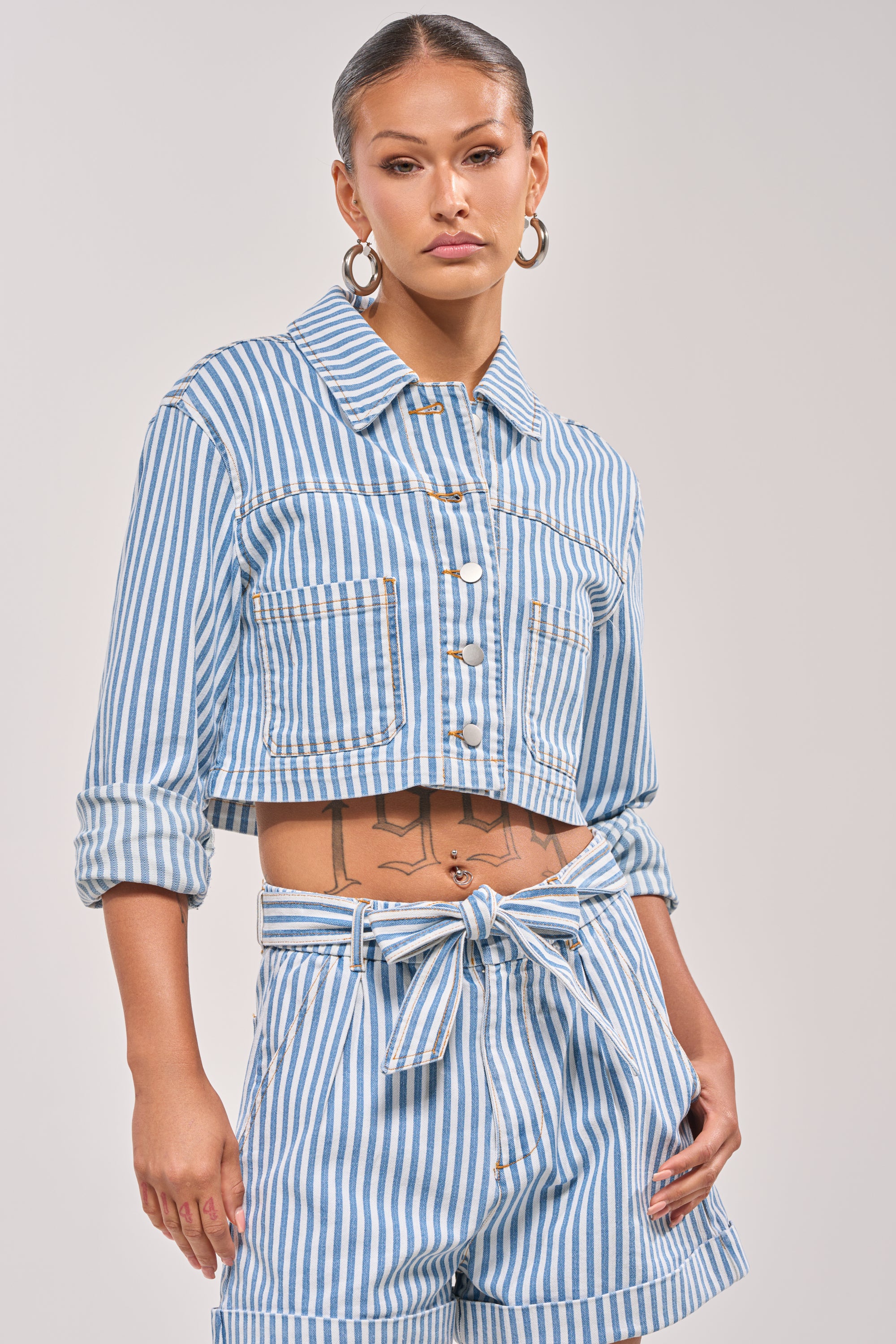 She wears the VACA VIBES BUTTON DOWN, a blue and white cropped boxy fit jacket paired with matching high-waisted tie-belt shorts, accessorized with hoop earrings and showcasing a torso tattoo for a bold resort-ready look against a light backdrop. 1