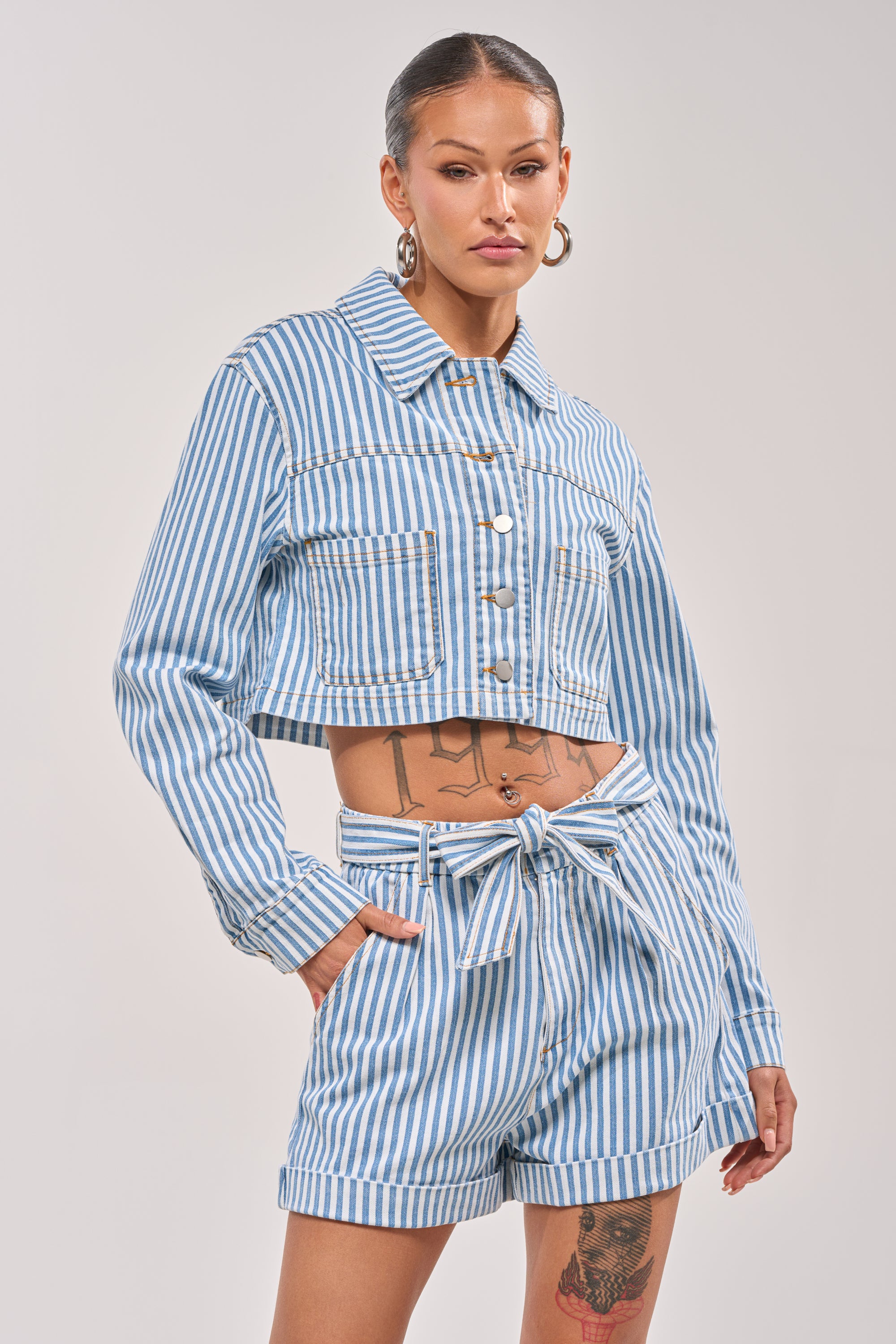 A woman models the VACA VIBES BUTTON DOWN, a blue and white vertical striped matching set with a cropped, boxy top and high-waisted tie-belt shorts. She accessorizes with hoop earrings, slicked-back hair, and visible tattoos. 2
