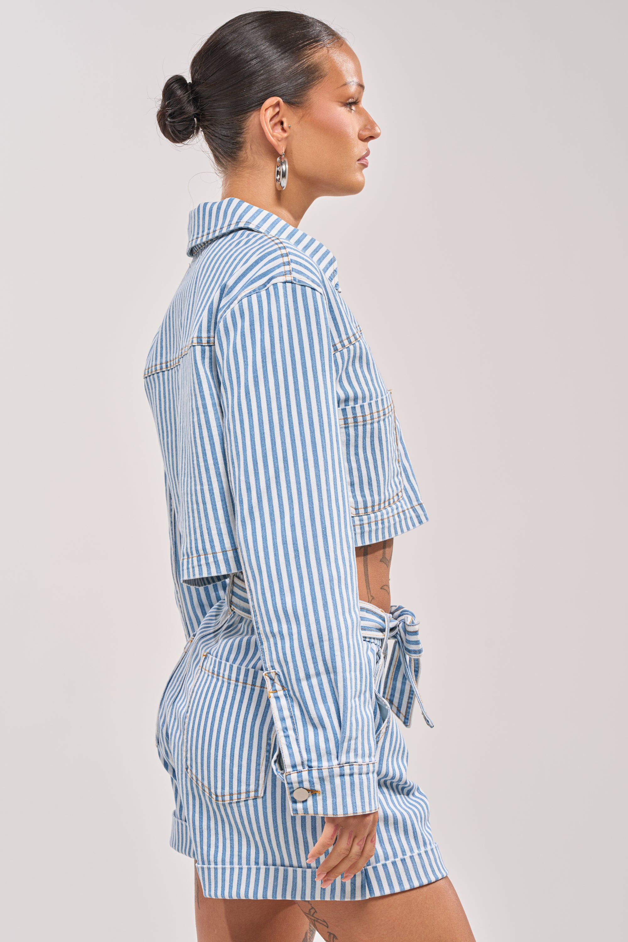 A woman with dark hair in a bun wears the VACA VIBES BUTTON DOWN, a blue and white striped, cropped boxy fit top, with matching high-waisted shorts, shown in profile against a plain background. 3