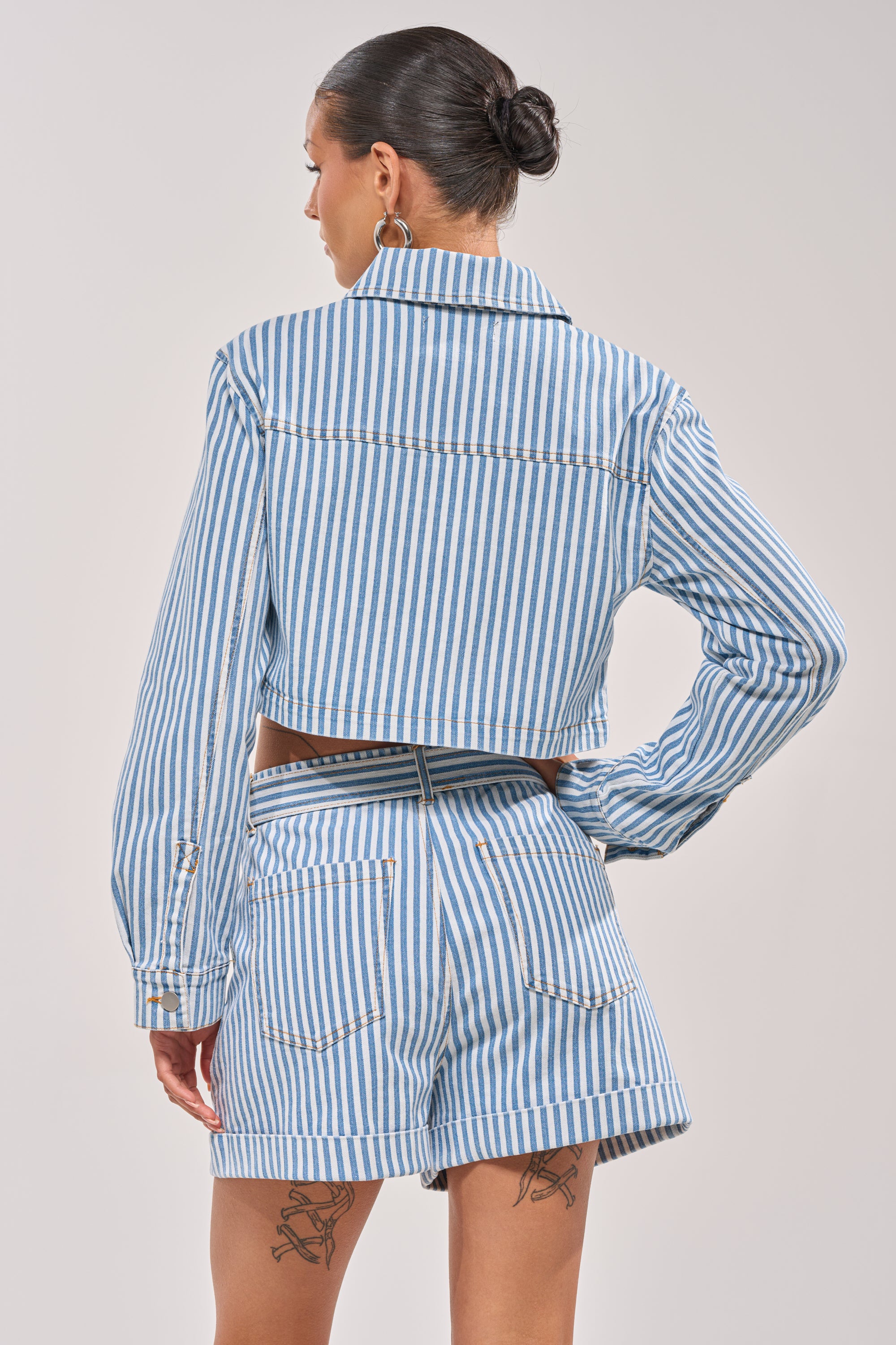 A woman with her hair in a bun wears the VACA VIBES BUTTON DOWN—a blue and white striped, cropped, boxy-fit top with matching high-waisted shorts. She stands with her back to the camera, showing two leg tattoos. 4