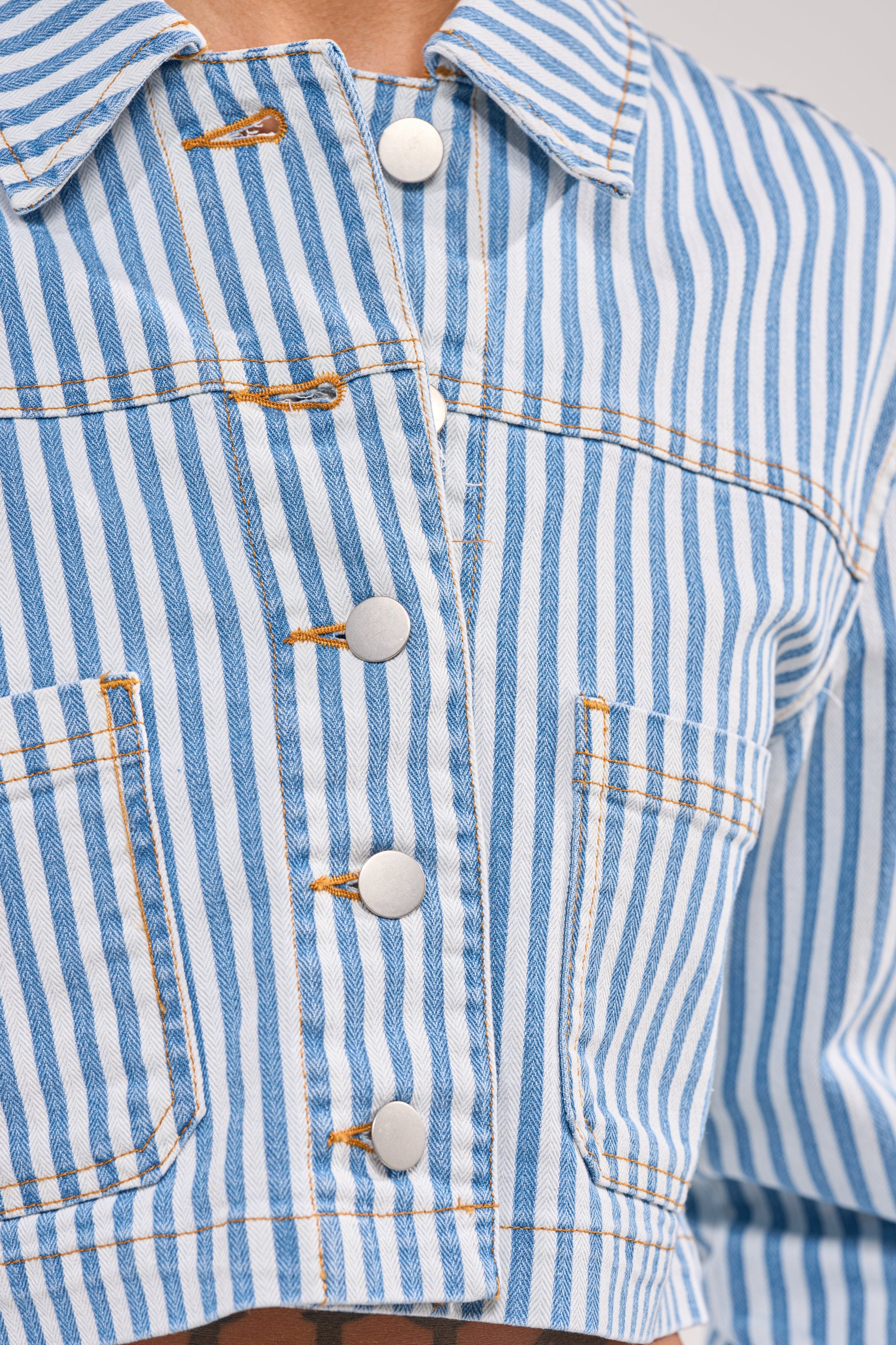 Close-up of the VACA VIBES BUTTON DOWN—a blue and white vertically striped, cropped boxy shirt with silver buttons, two front pockets, visible yellow stitching, and a resort-ready style. 7