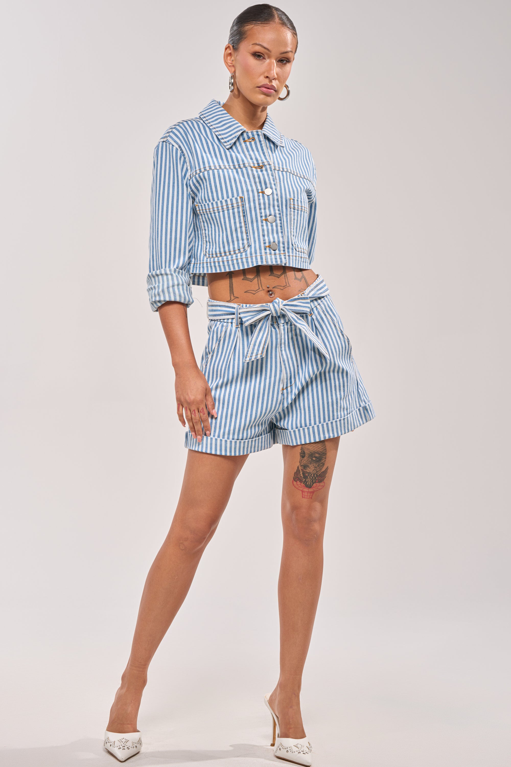 A woman poses in VACA VIBES SHORT, a blue and white nautical striped high-rise short with a tie belt, paired with a matching cropped jacket, white heels, hoop earrings, and shows off her leg and torso tattoos. 0