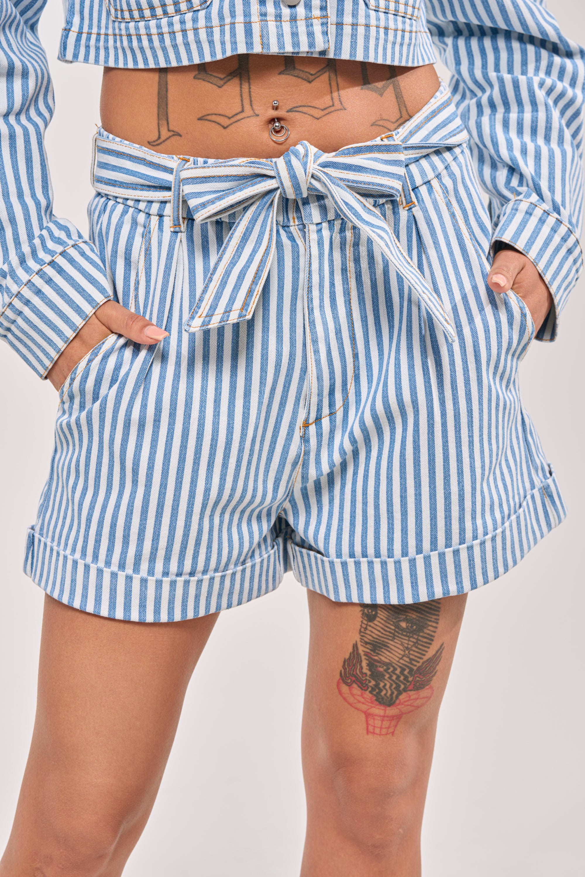Wearing the VACA VIBES SHORT—blue and white nautical striped high-rise shorts with a matching belt—this person pairs it with a coordinating striped top, hands in pockets, showing visible tattoos on the torso and thigh. 1