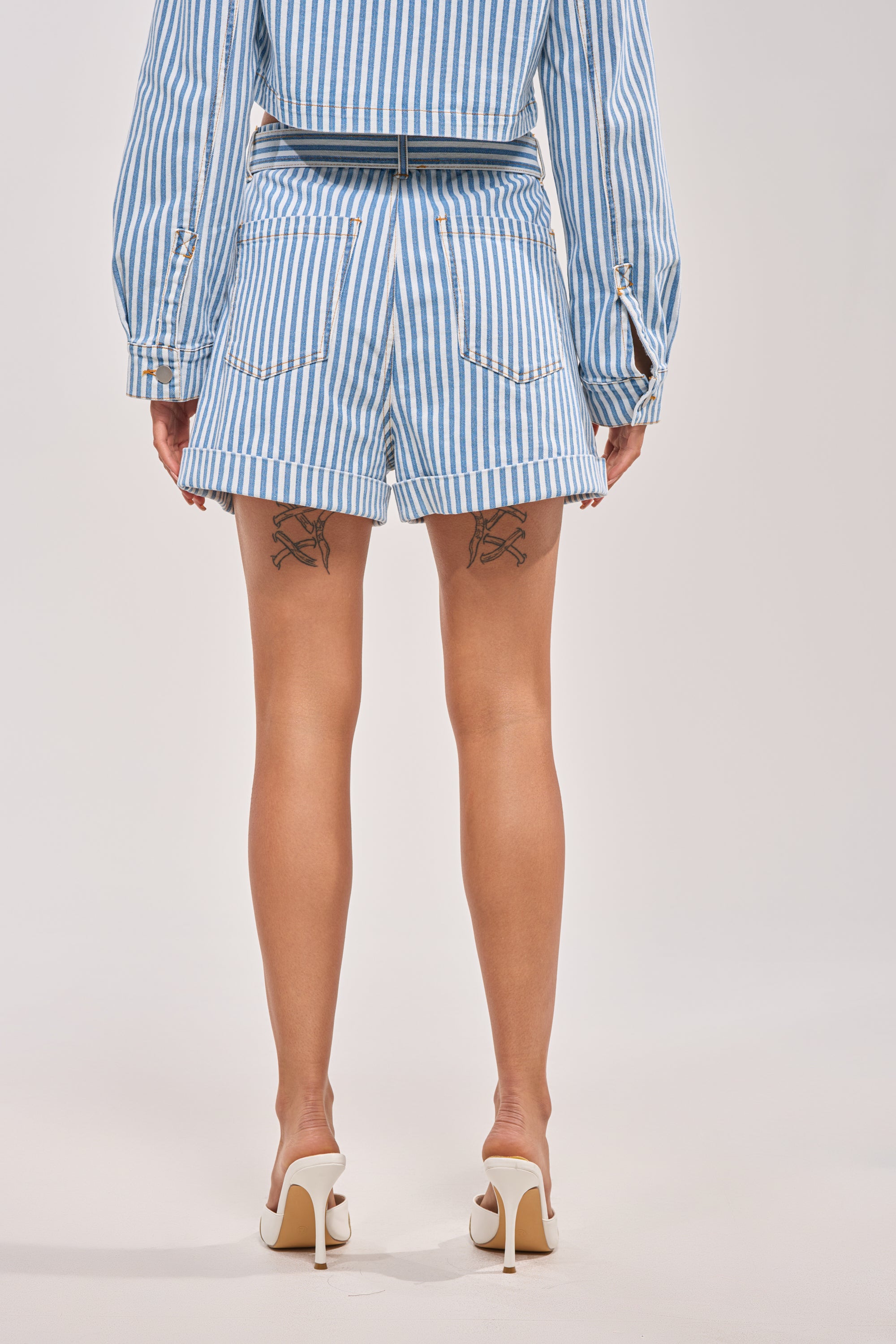 A person stands with their back to the camera, wearing the VACA VIBES SHORT in blue and white nautical stripes, paired with a matching jacket. Thigh tattoos and white high heels complete this striking look. 5