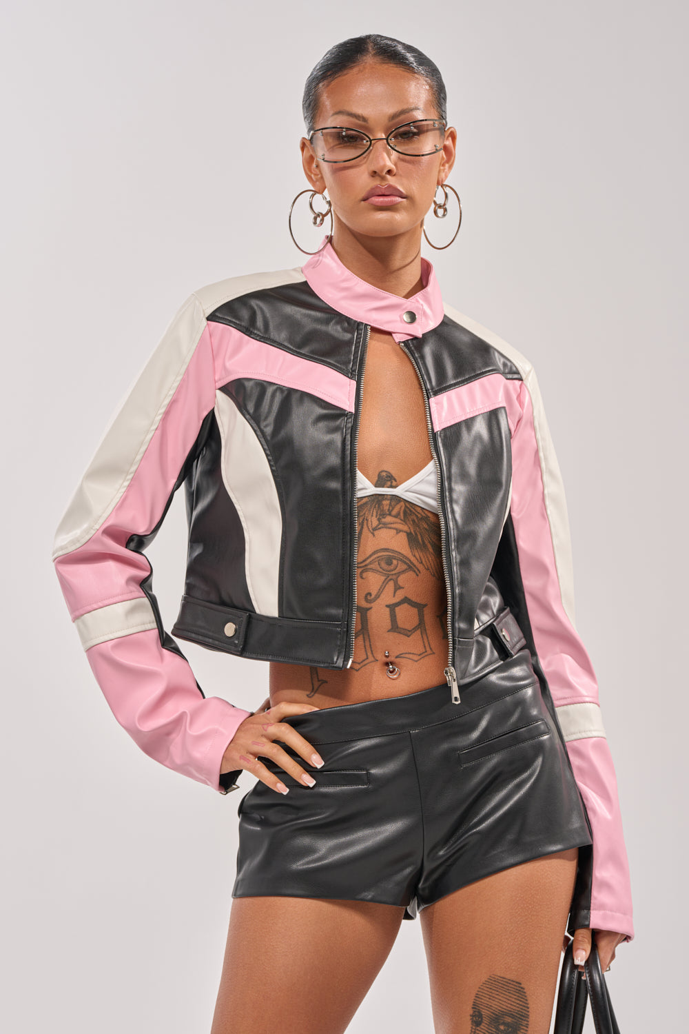 A confident woman showcases bold streetwear in the LILOU EVERYDAY MOTO color-block jacket, black faux leather shorts, hoop earrings, sunglasses, and a handbag. Tattoos on her torso and thigh complete the striking look. 0