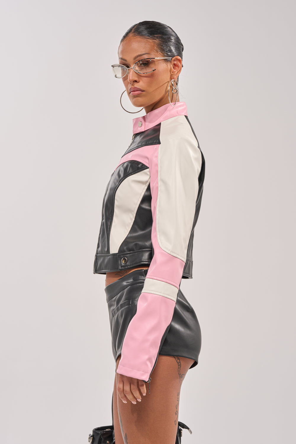 A woman in the LILOU EVERYDAY MOTO—a color-block faux leather jacket in black, white, and pink—paired with matching shorts, hoop earrings, and stylish glasses stands in profile against a plain background. 3