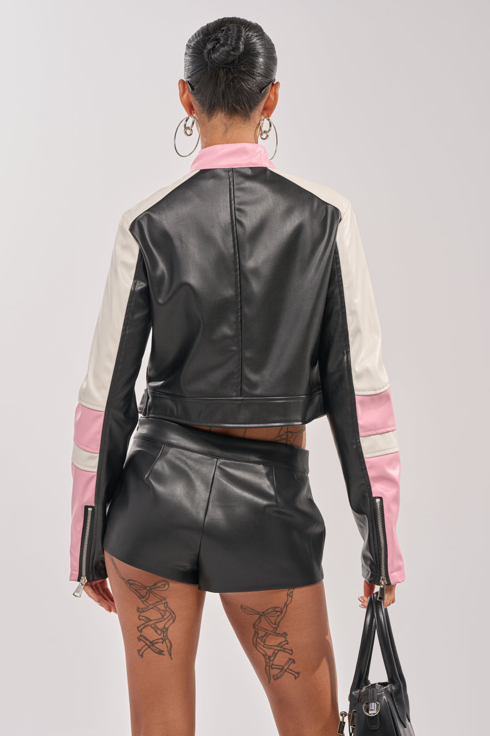 A woman stands facing away, wearing the LILOU EVERYDAY MOTO jacket with white and pink sleeves, matching black faux leather shorts, hoop earrings, visible leg tattoos, and holding a small black handbag. 4