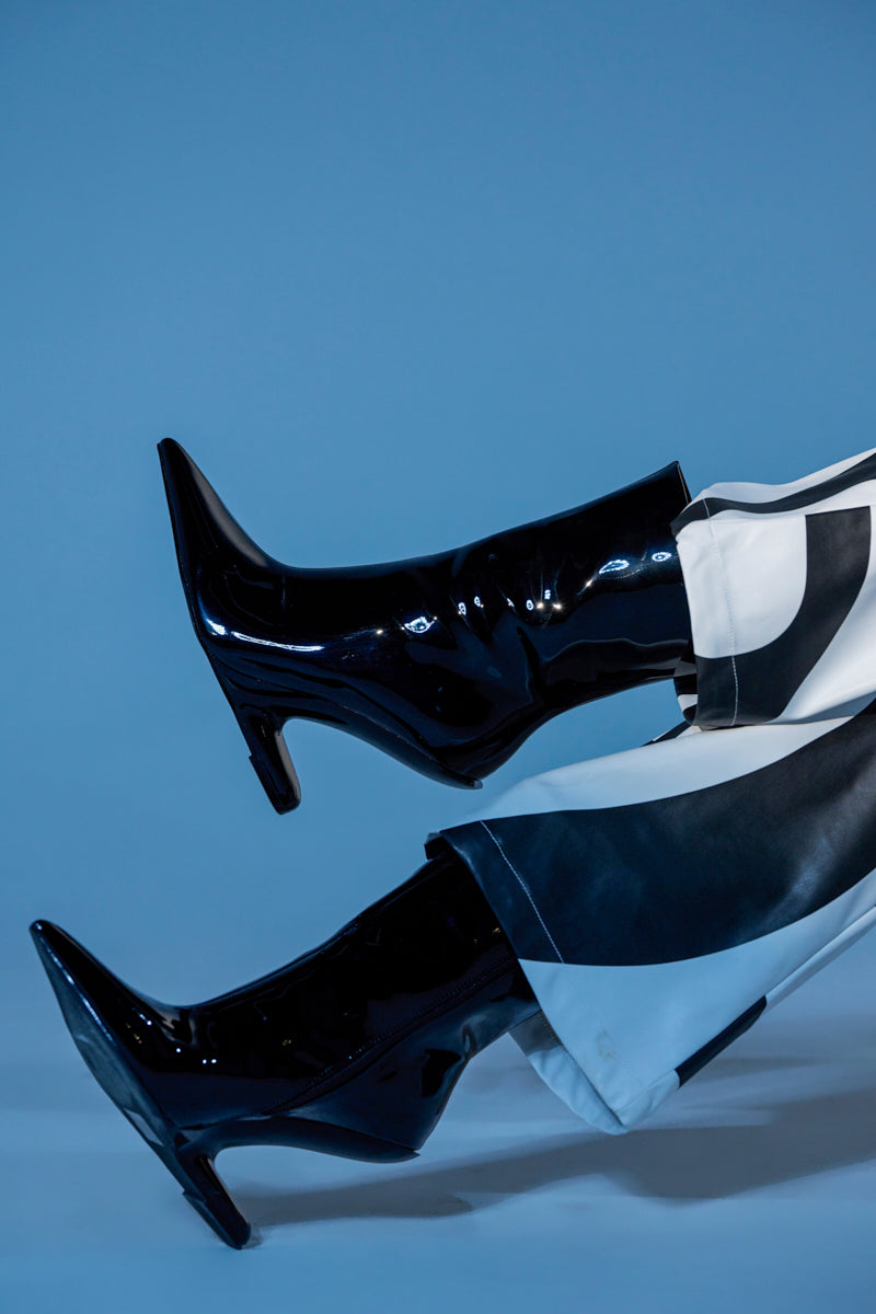 A person wearing AZALEA WANG GLACIELLE PATENT BLACK WEDGE BOOTIE heels and black-and-white patterned pants poses with legs raised against a plain blue background.