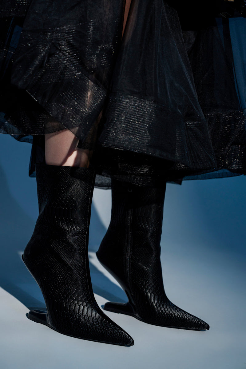A person stands on a light blue surface wearing the AZALEA WANG GLACIELLE BLACK CROC BOOTIE, which features pointed toes and unique inverted heels, paired with a sheer black skirt.