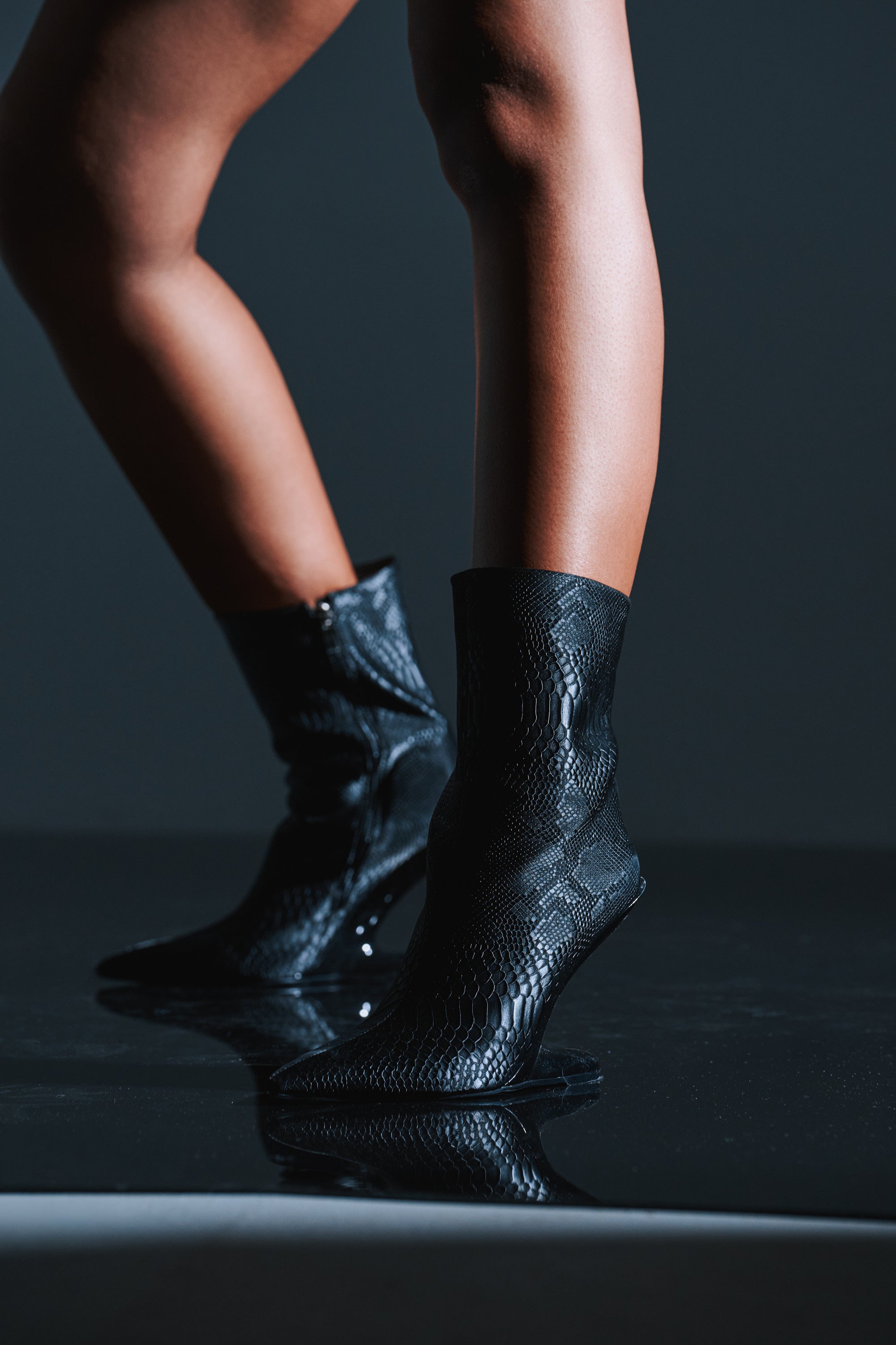 Someone stands on a glossy, reflective surface against a dark background, wearing the AZALEA WANG GLACIELLE BLACK CROC BOOTIE with a pointed toe and snakeskin pattern.
