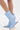 Someone wears the AZALEA WANG GLACIELLE BLUE WEDGE BOOTIE—a mid-calf, light blue velvet boot with a pointed toe and sculptural wedge heel—while standing against a white background.