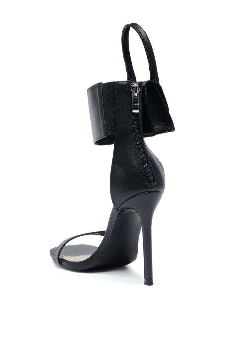 The AZALEA WANG GLENDA BLACK BAG EMBELLISHMENT SANDAL features a black faux leather upper, open toe, slender stiletto heel, wide ankle strap with back zipper, and bag embellishment. Photographed against a white background. 6