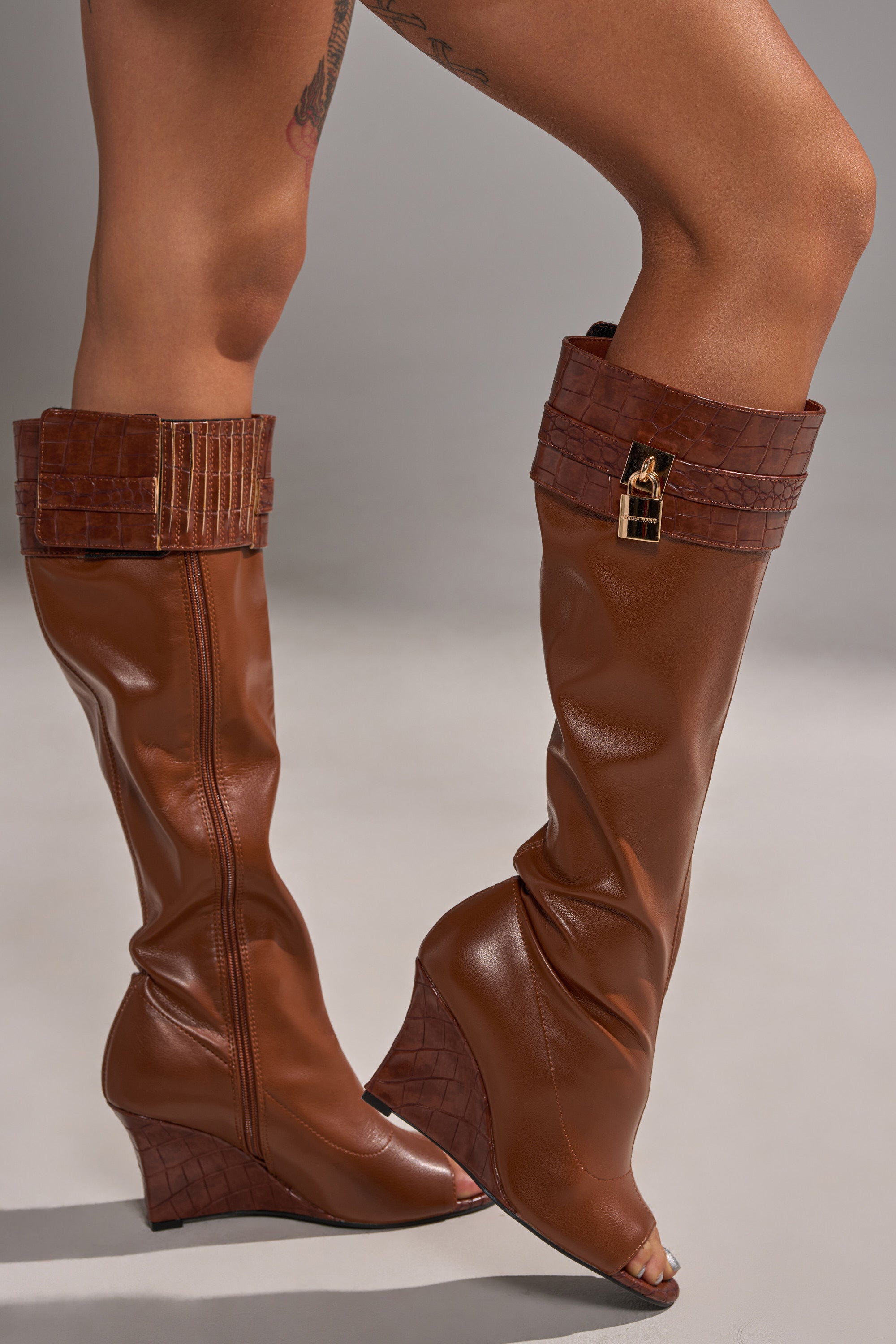 A person wears the AZALEA WANG GLENNA COGNAC STRETCH PU WEDGE BOOT, featuring knee-high length, crocodile-textured cuff, gold padlock detail, and open toes, standing with one leg bent and showing leg tattoos.