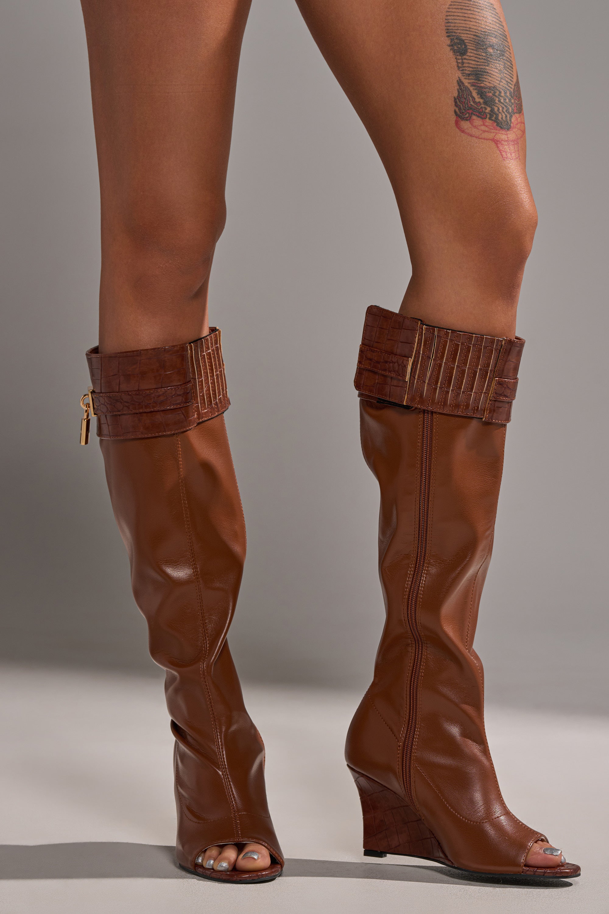 A person wears AZALEA WANG GLENNA COGNAC STRETCH PU WEDGE BOOTS with cuffed tops and gold zipper detail. Their bare legs reveal a skull tattoo on the right thigh. The background is neutral gray.