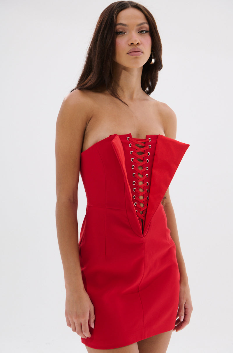 A woman wears the CHAUNTELLE LACE UP MINI DRESS, a strapless, fitted red mini dress featuring a plunging neckline and lace-up grommet details down the front. She has long straight hair and stands against a plain white background.
