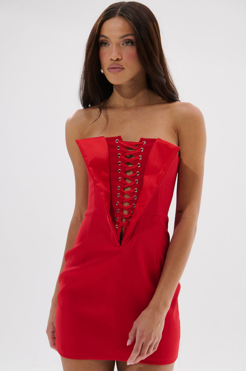 A woman with long brown hair models the CHAUNTELLE LACE UP MINI DRESS, a strapless red mini dress featuring a corset-style lace-up front with grommet details. She stands against a plain white background, gazing to the side.