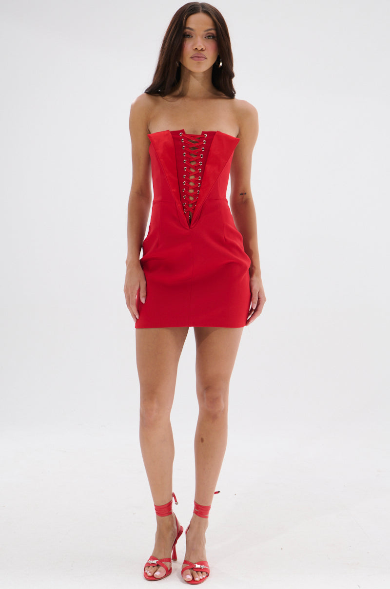 A woman stands against a white background wearing the CHAUNTELLE LACE UP MINI DRESS in red, featuring a strapless design, lace-up front, fitted corset-style detailing, and matching red high-heeled sandals.