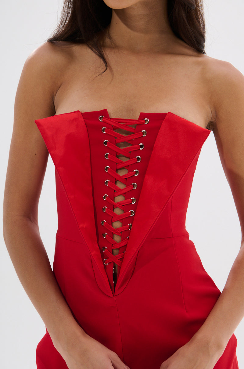 A woman models the CHAUNTELLE LACE UP MINI DRESS, a red strapless corset-style mini with a plunging neckline, silver eyelets, and black lace-up detailing down the front. Her arms rest at her sides as she turns her head slightly.