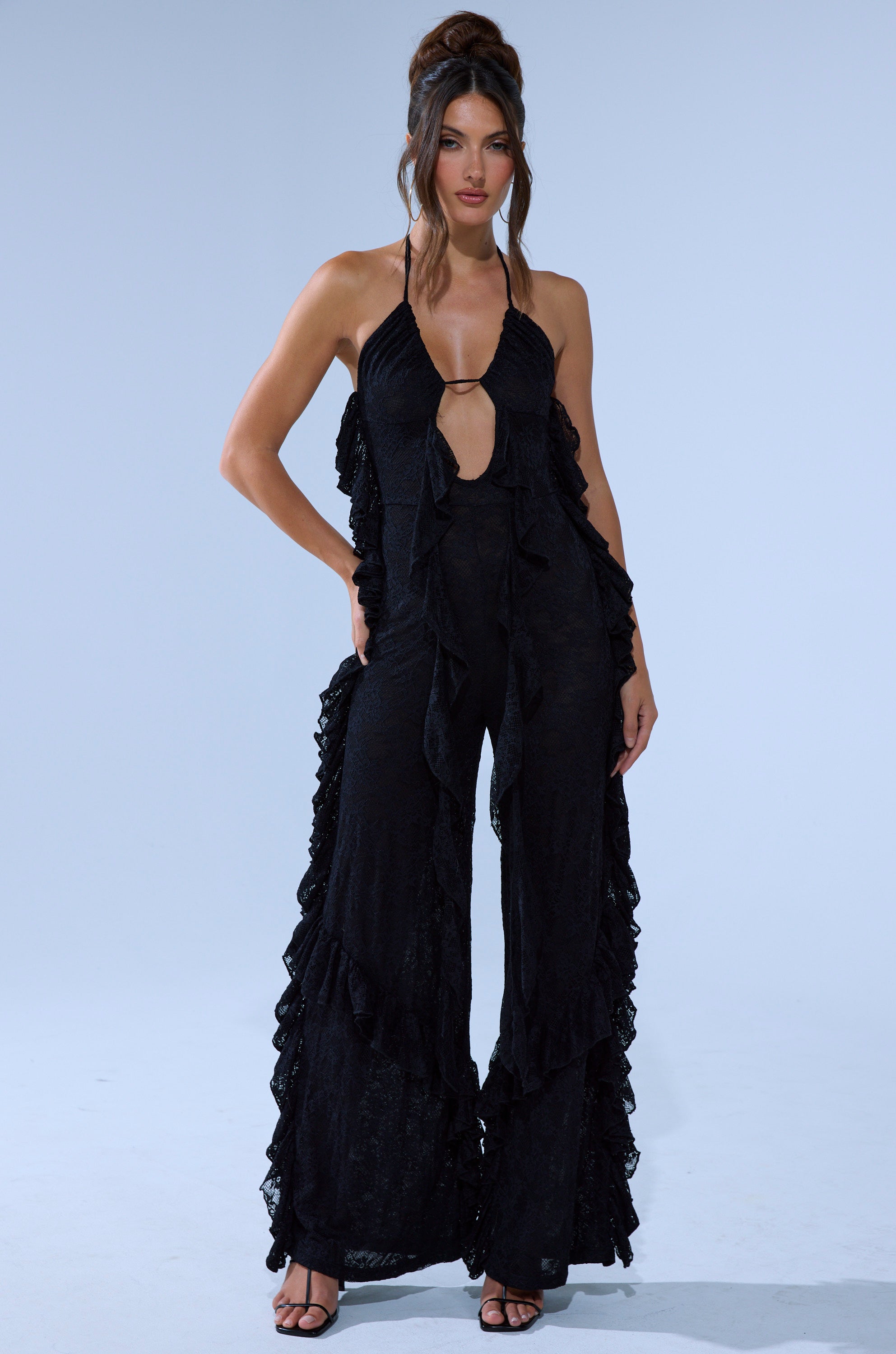 A woman wears the FREE AS A BIRD RUFFLED JUMPSUIT, a sleeveless black lace one-piece featuring ruffle details on the legs and neckline. Her long hair is styled up, and she pairs the look with black high-heeled sandals against a plain background.