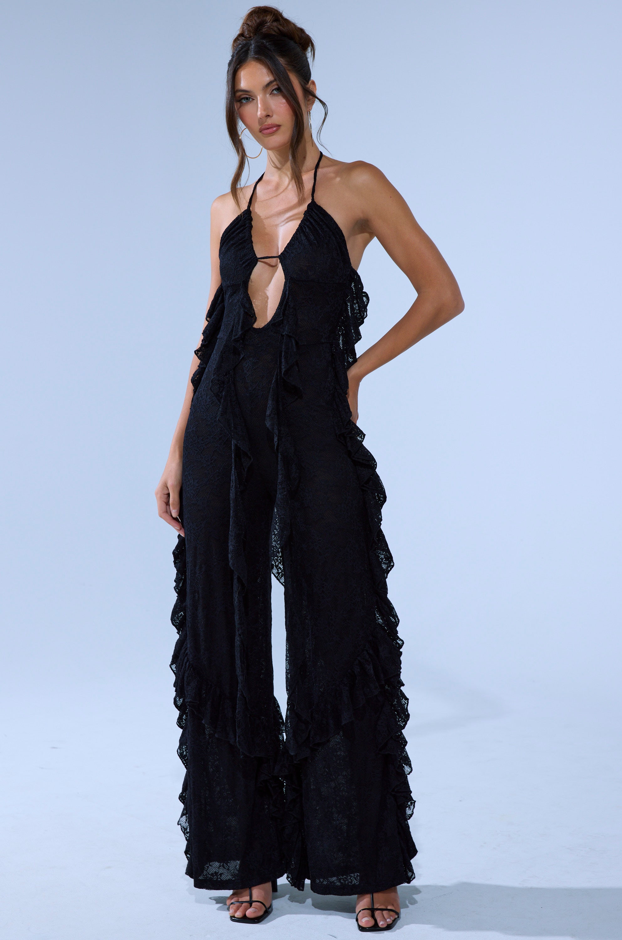 A woman poses against a light gray background in the FREE AS A BIRD RUFFLED JUMPSUIT, featuring black lace, ruffles, and a wide-leg silhouette. Her long wavy brown hair is tied up, and she wears black high-heeled sandals.