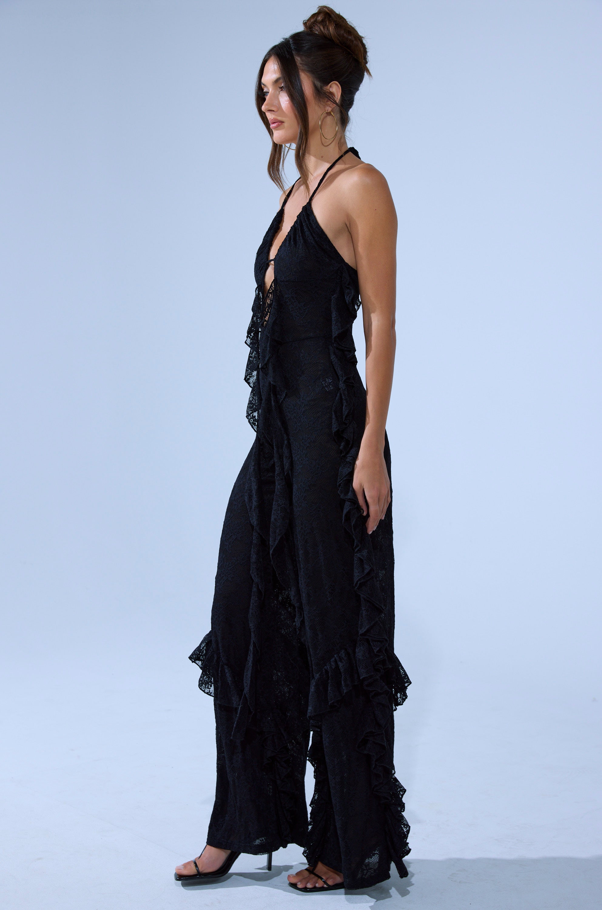 A woman in the FREE AS A BIRD RUFFLED JUMPSUIT, featuring a black, sleeveless, wide-leg silhouette and ruffled details, stands in profile with strappy heels and large hoop earrings against a plain light background. Her hair is in a messy bun.