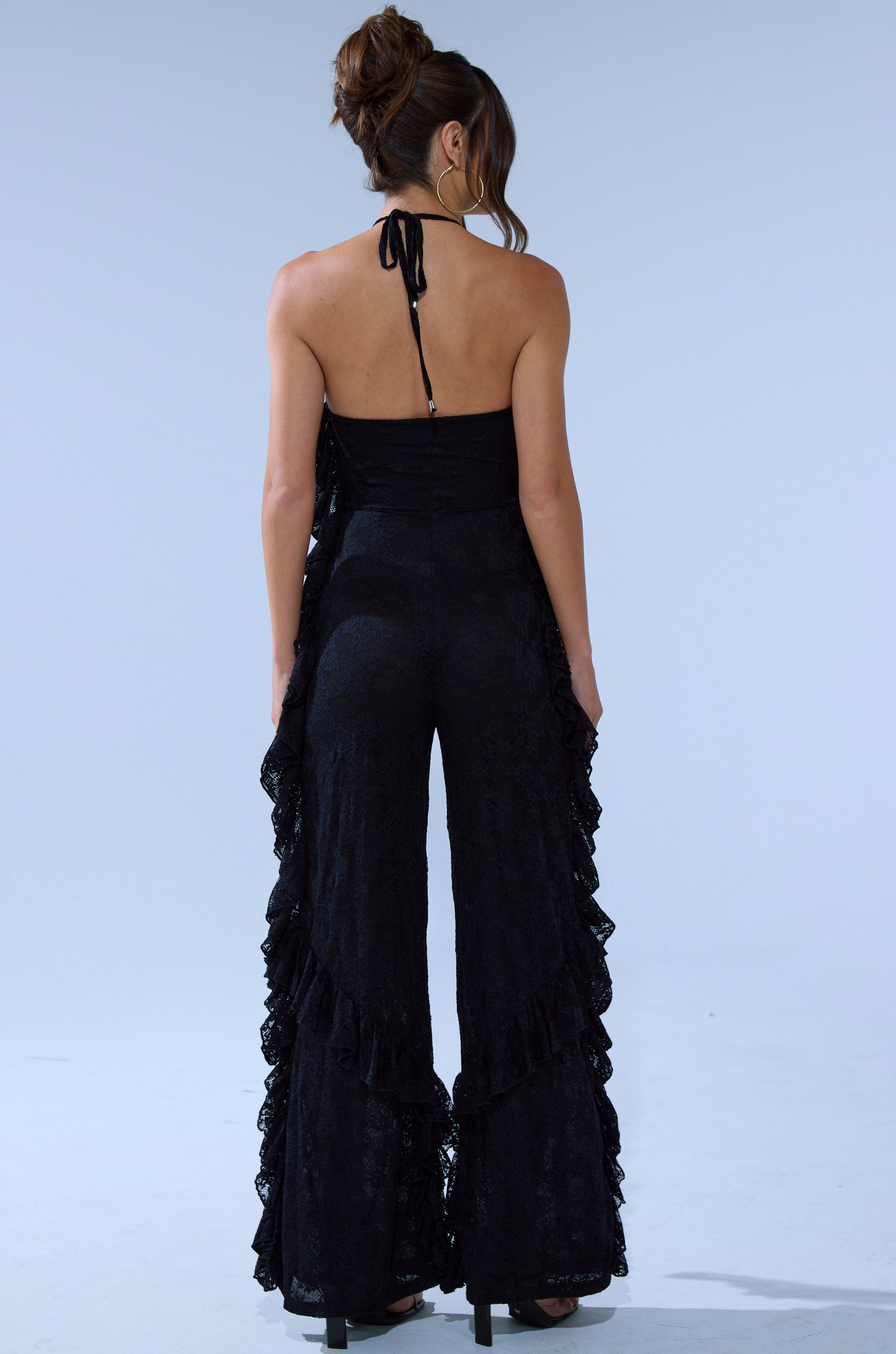 A woman with her hair in a messy bun poses against a plain light background, facing away and wearing the FREE AS A BIRD RUFFLED JUMPSUIT featuring halter straps and flared, ruffled pant legs.