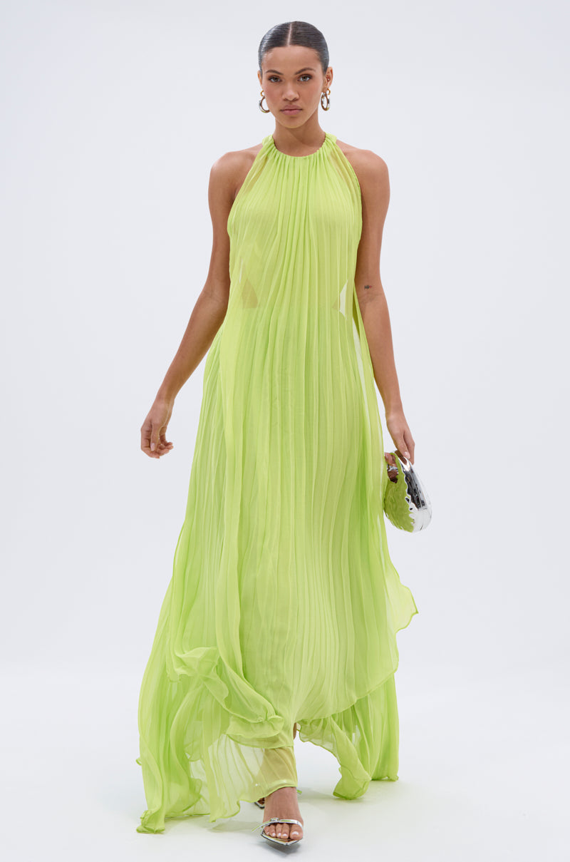 A woman strides confidently in the NIXEN MAXI DRESS WITH CHIFFON OVERLAY, a flowing, pleated neon green dress. She carries a small clutch, wears hoop earrings and strappy heels against a plain white background.