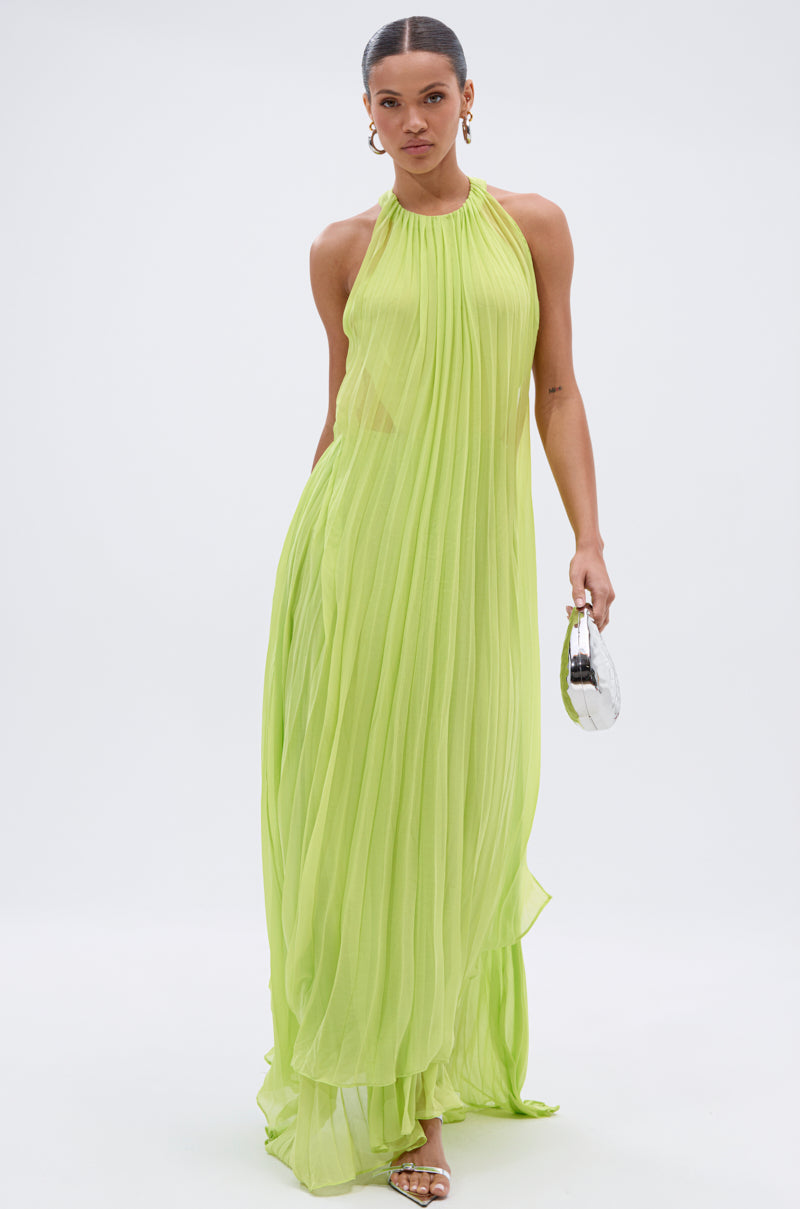 A woman in the NIXEN MAXI DRESS WITH CHIFFON OVERLAY, a sheer neon green maxi, stands against a white background holding a silver clutch and wearing gold hoop earrings and sandals.
