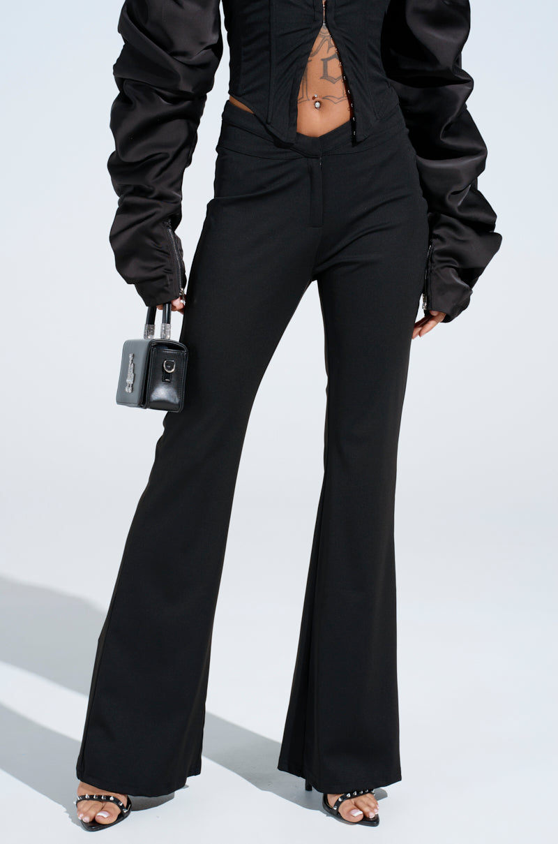 A person in black FEELING EXTRA FLARE PANTs, an off-shoulder top with puffy sleeves, and open-toed heels holds a small black handbag against a light gray background.