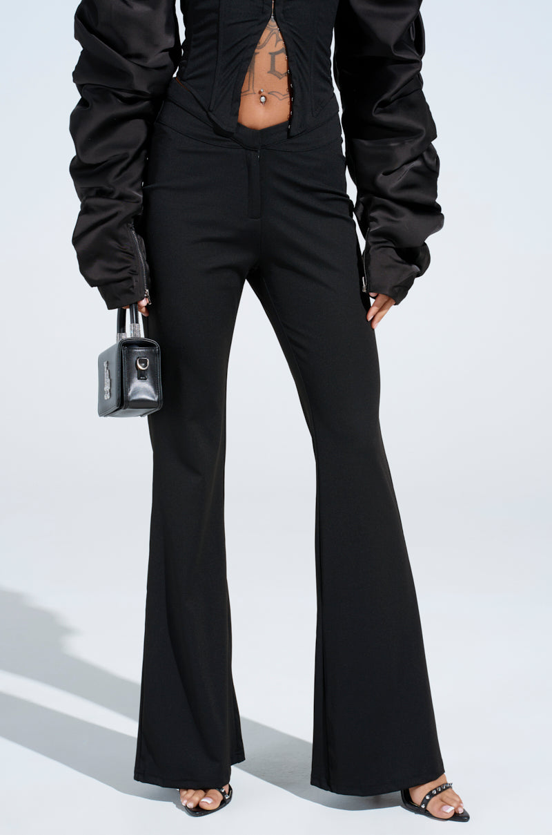 Dressed in a chic black ensemble featuring the FEELING EXTRA FLARE PANT and a ruched long-sleeve top, a person holds a small black handbag and wears open-toed sandals. The plain, light-colored background enhances the stylish look.