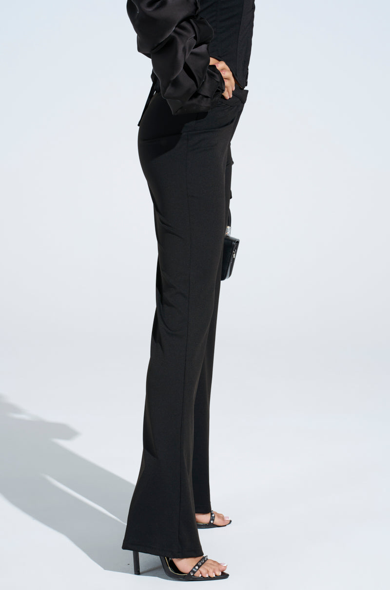 Dressed in FEELING EXTRA FLARE PANT with a mid-rise fit and slight flare, the individual stands sideways against a plain white background. Their ensemble includes a curve-sculpting satin top, black high-heeled sandals, and they're holding a small black purse.
