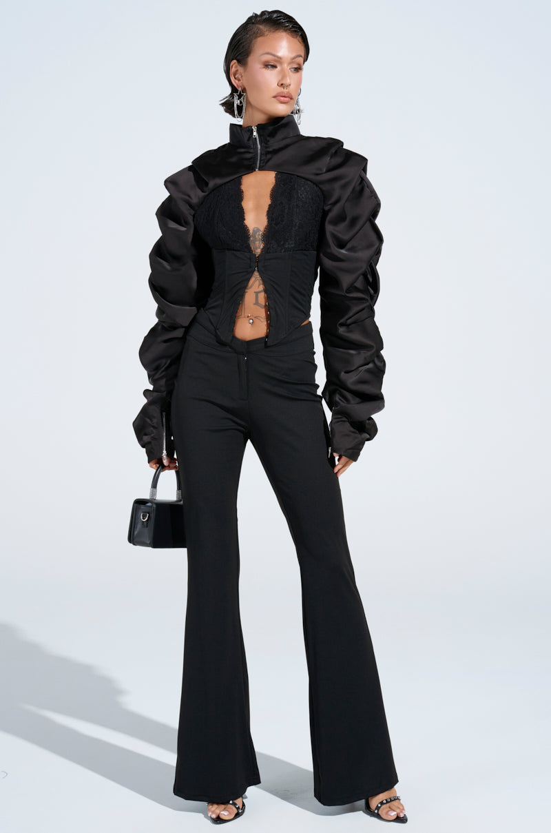 Dressed in a chic black ensemble, the person wears a fitted jacket with voluminous sleeves and corset-style front, FEELING EXTRA FLARE PANTs that enhance curves, a small black handbag, and open-toe shoes against a simple backdrop.