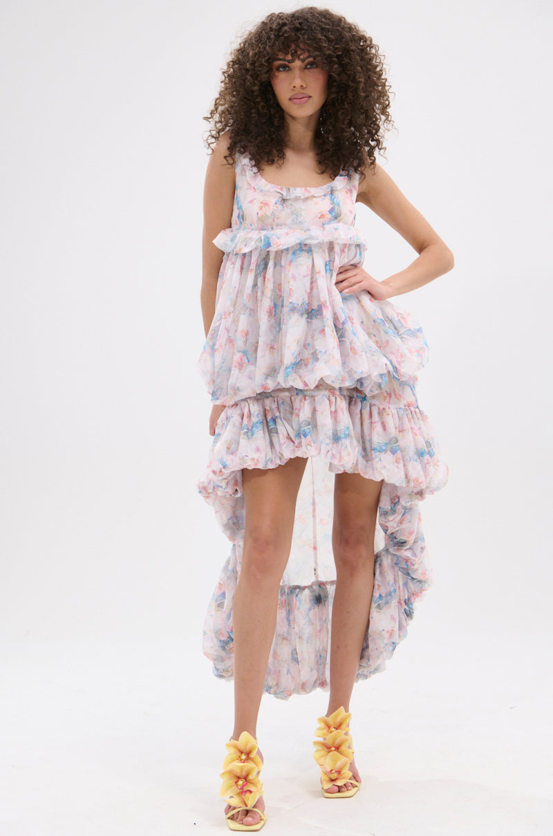 A woman with curly hair confidently poses in the THROUGH THE MEADOW RUFFLE HIGH LOW DRESS, featuring a ruffle hemline and pastel floral pattern, styled with yellow ruffled high-heeled sandals against a plain white background.