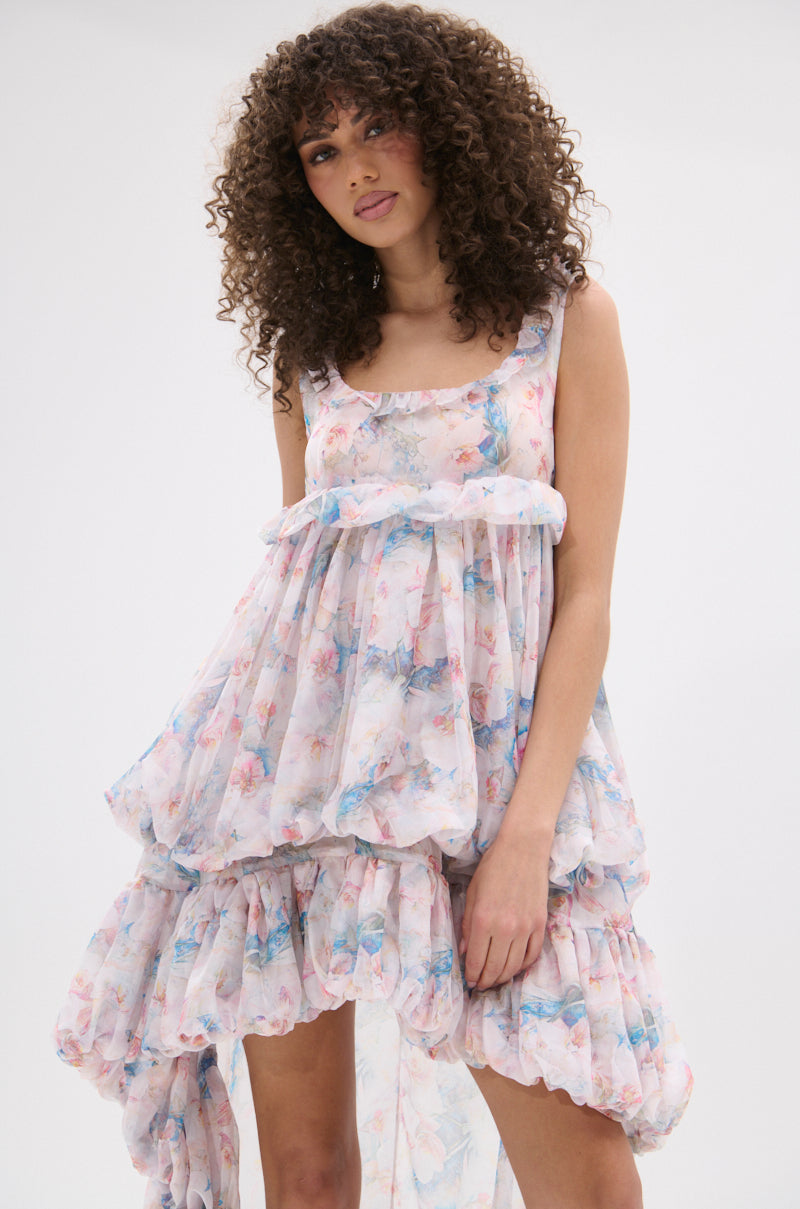 A woman with curly hair models the THROUGH THE MEADOW RUFFLE HIGH LOW DRESS, a sleeveless, flowy pastel floral dress with ruffled layers and a high-low hem, set against a plain white background.