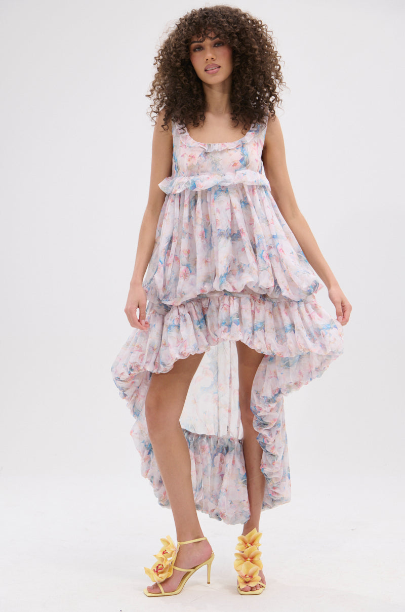 A woman with curly hair wears the THROUGH THE MEADOW RUFFLE HIGH LOW DRESS, a pastel floral high-low dress with a ruffle hem, and yellow flower-detailed heels, standing against a plain white background.