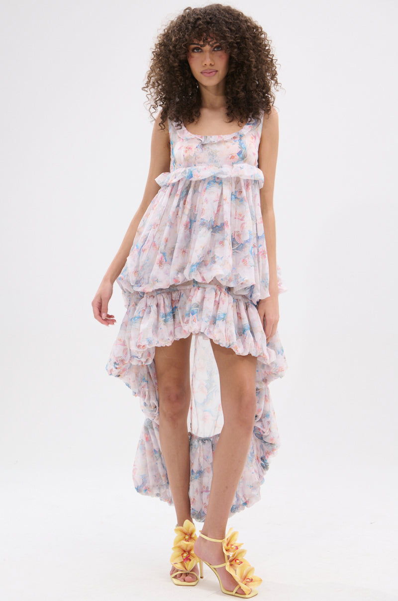 A woman with curly hair wears the THROUGH THE MEADOW RUFFLE HIGH LOW DRESS and yellow ruffled heels, standing against a plain white background.