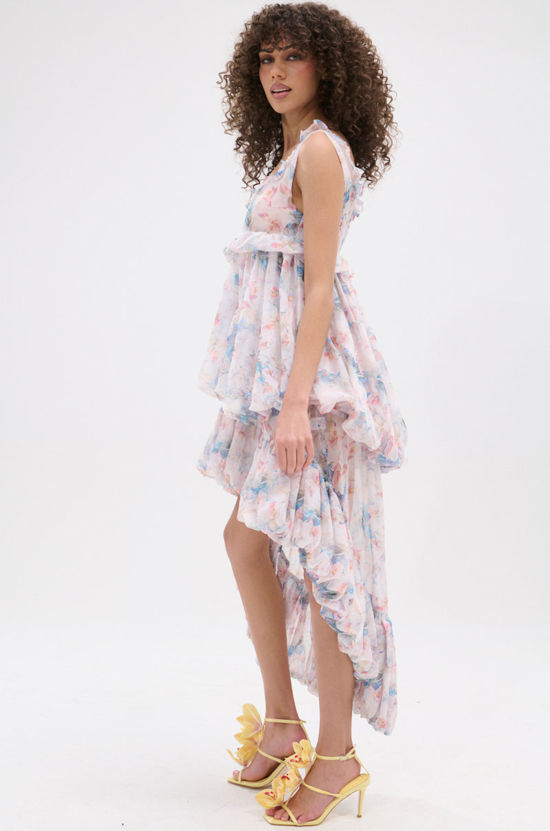 A woman with curly hair models the THROUGH THE MEADOW RUFFLE HIGH LOW DRESS—pastel floral, sleeveless, and high-low hem—paired with yellow leaf-detailed heeled sandals, standing sideways against a plain white background.